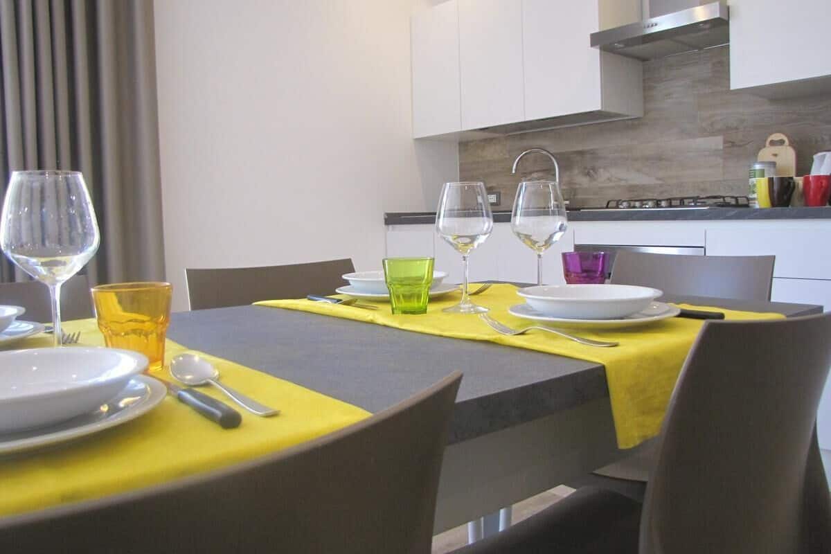 A modern dining area with a table set for two, featuring a yellow tablecloth, wine glasses, and a green glass.