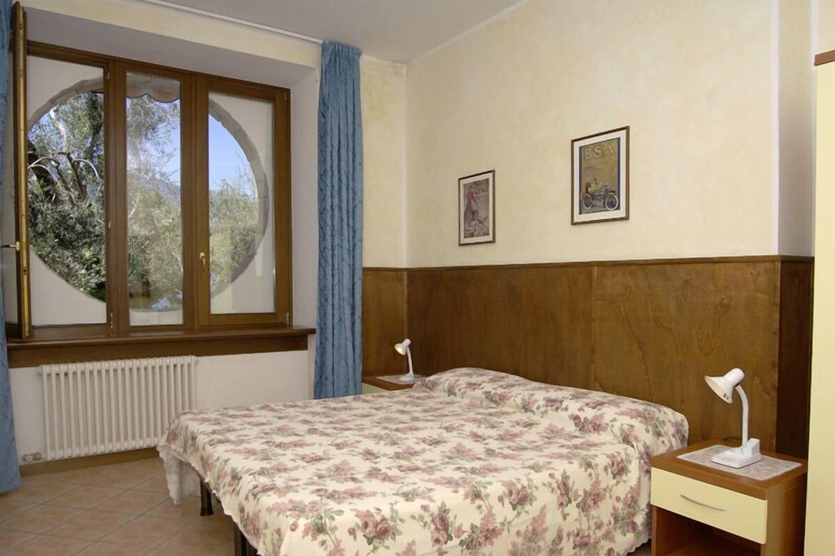 A bedroom with a bed, two bedside tables, a window with a view of trees, and two framed pictures on the wall.