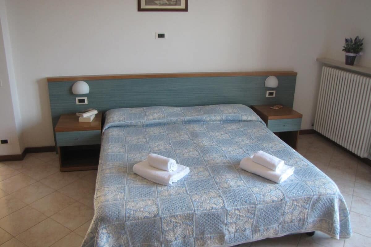 A neatly made bed with a patterned bedspread and two folded towels on the side. There are two bedside tables with lamps and a small picture frame on the wall above the bed.