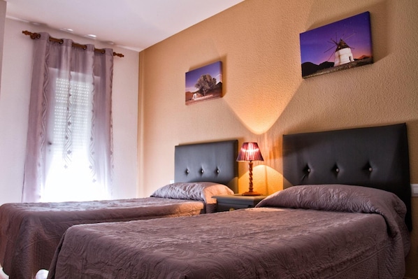 A hotel room with two beds, a nightstand, a lamp, and wall art depicting a windmill and a tree.