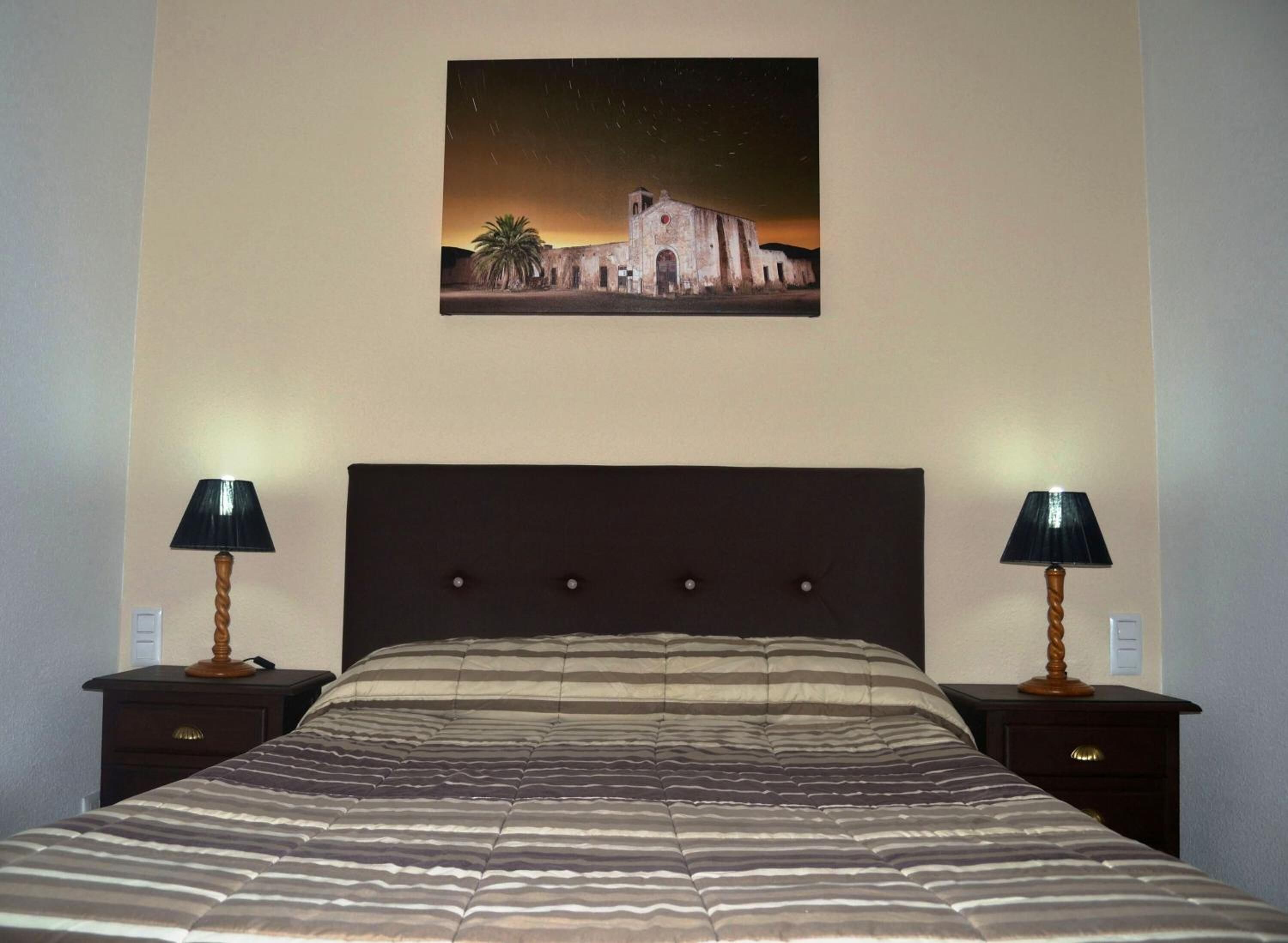 Una cama con colcha a rayas, dos mesitas de noche con lámparas, y un cuadro de un edificio en la pared.