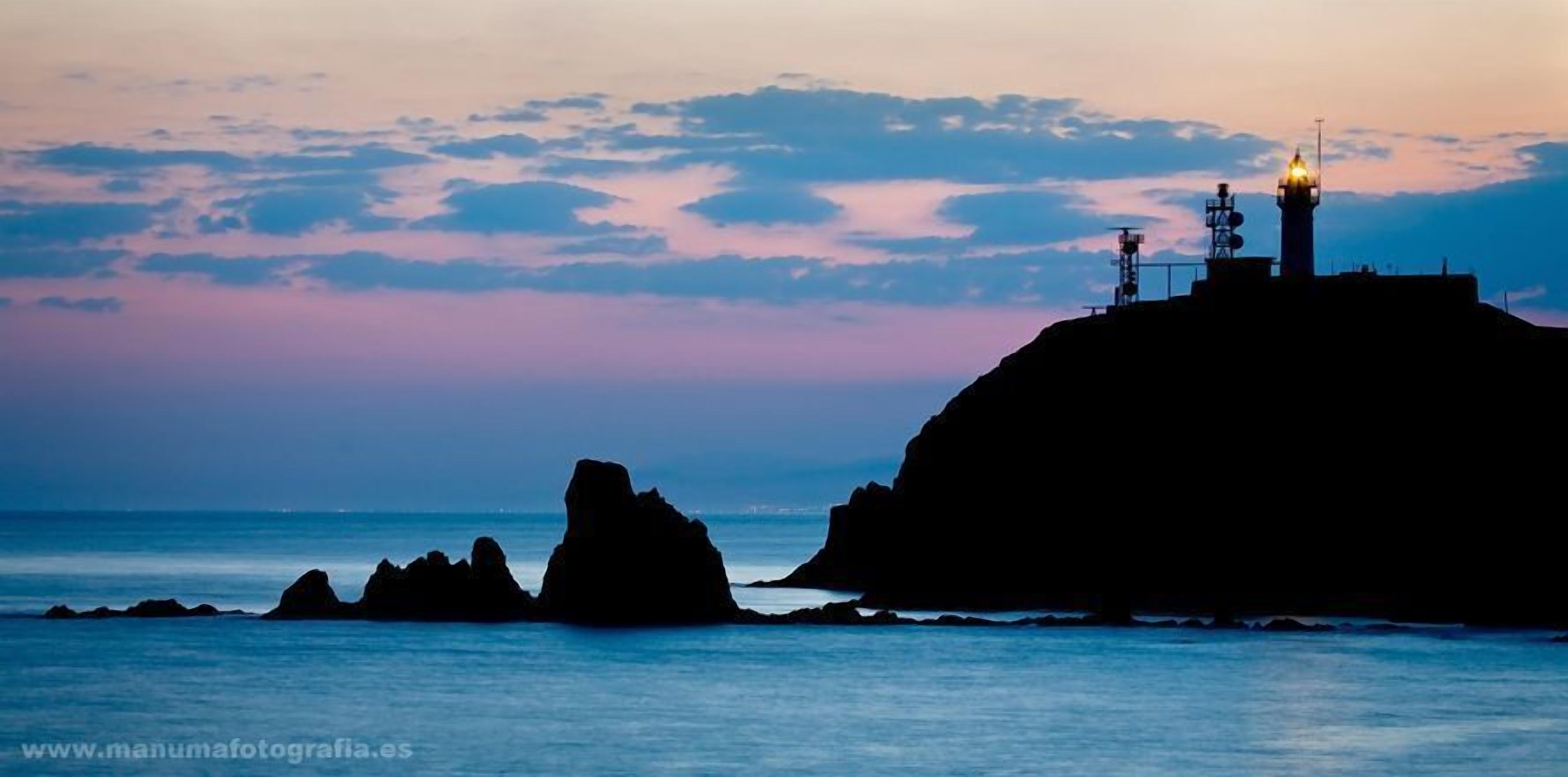 A coastal scene with a lighthouse on a cliff, calm sea, and a clear sky.