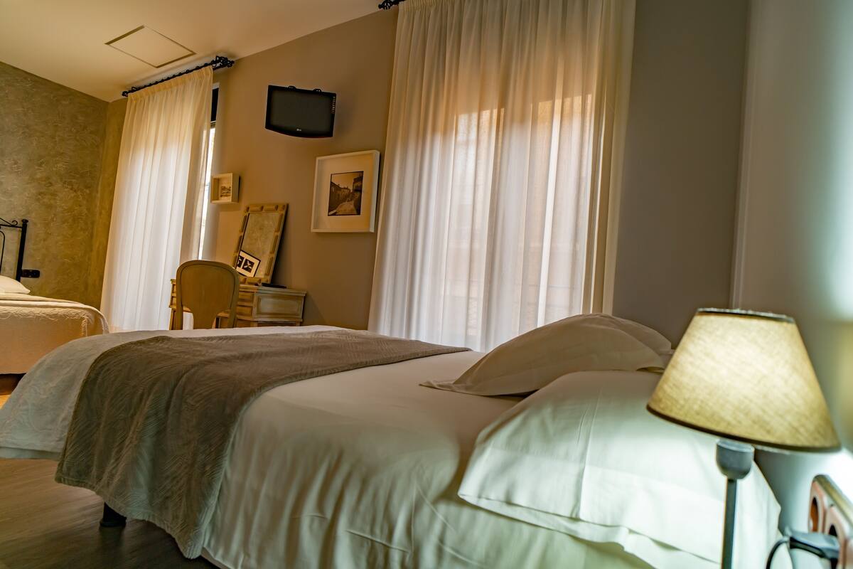 A hotel room with two beds, a bedside table, a lamp, a television on the wall, and a window with curtains.
