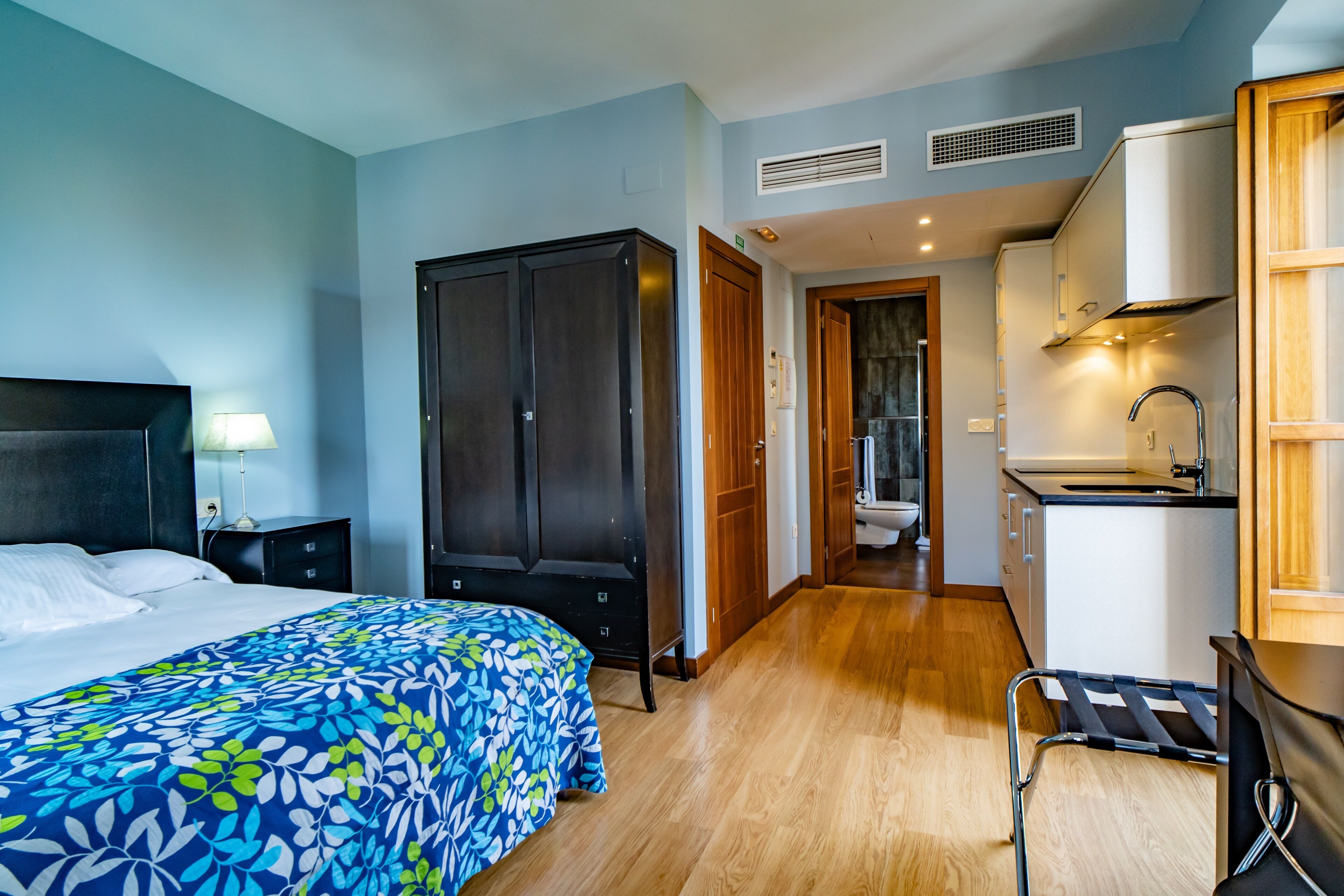 A hotel room with a bed, a wardrobe, a desk with a chair, a sink, and a view of the bathroom.