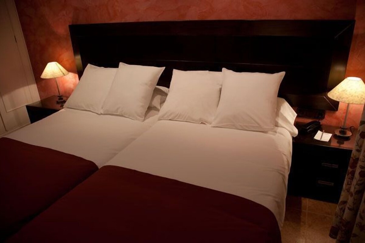 Uma cama de casal com lençóis brancos e duas almofadas, com mesas de cabeceira com luminárias de cada lado, em um quarto com cabeceira escura e parede em tons quentes.
