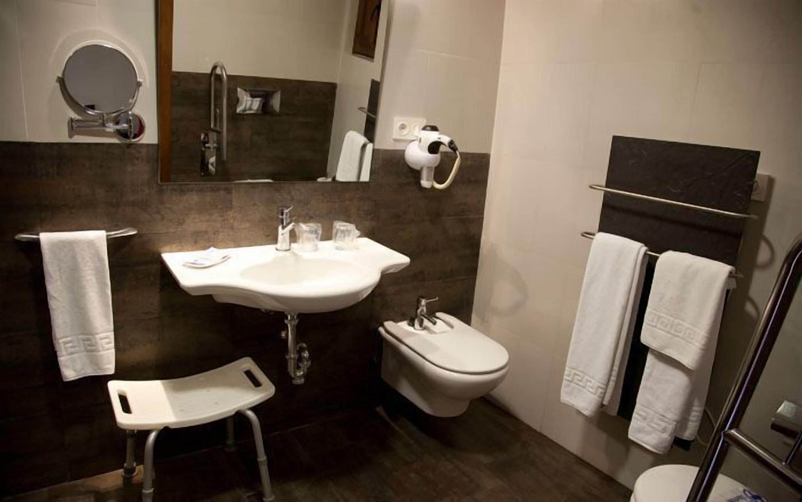 Un baño moderno con lavamanos, inodoro y bidé. Hay un taburete pequeño y un perchero para toallas.