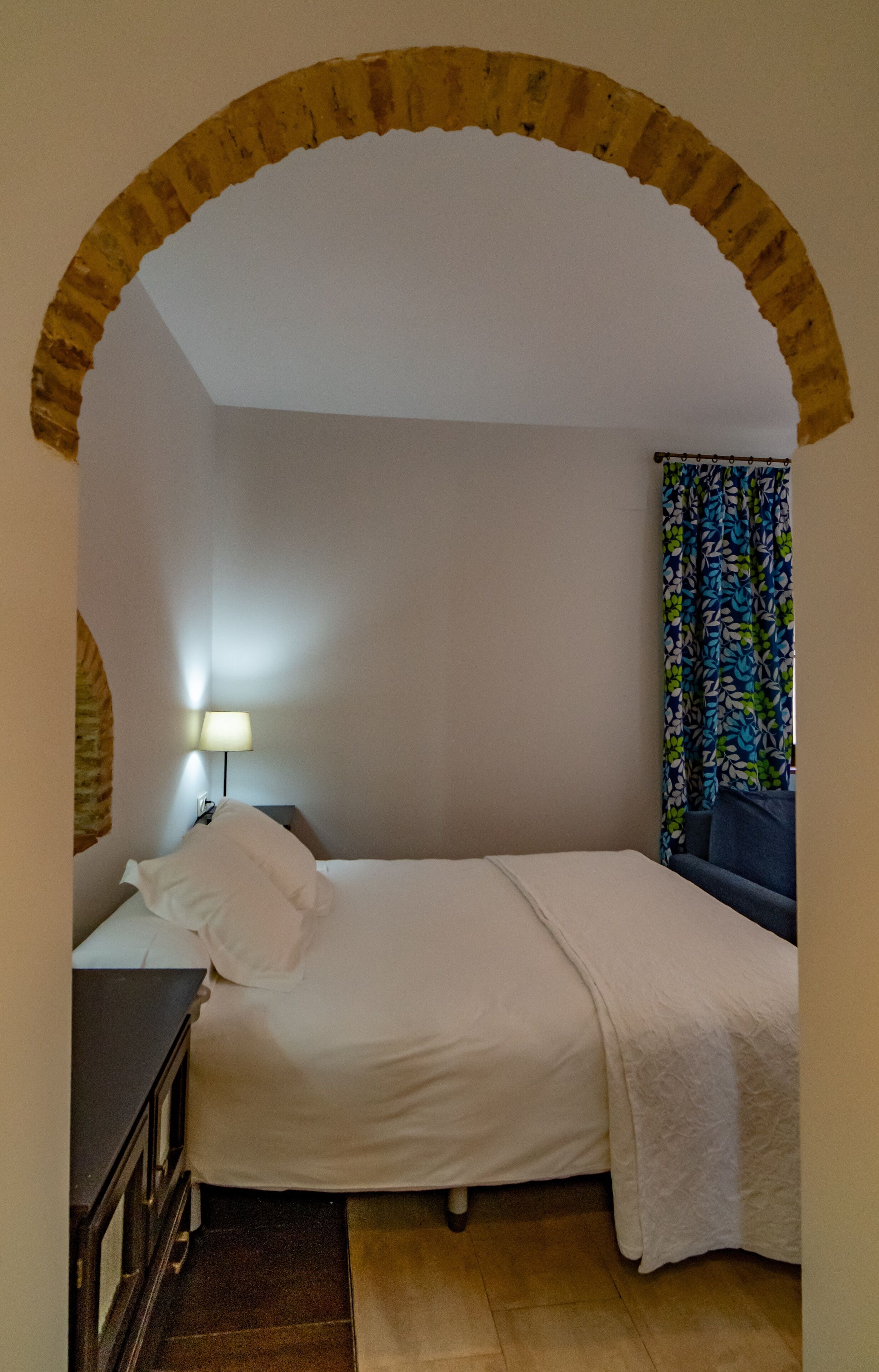 Un dormitorio con cama, mesita de noche, lámpara, una silla azul y cortina estampada.