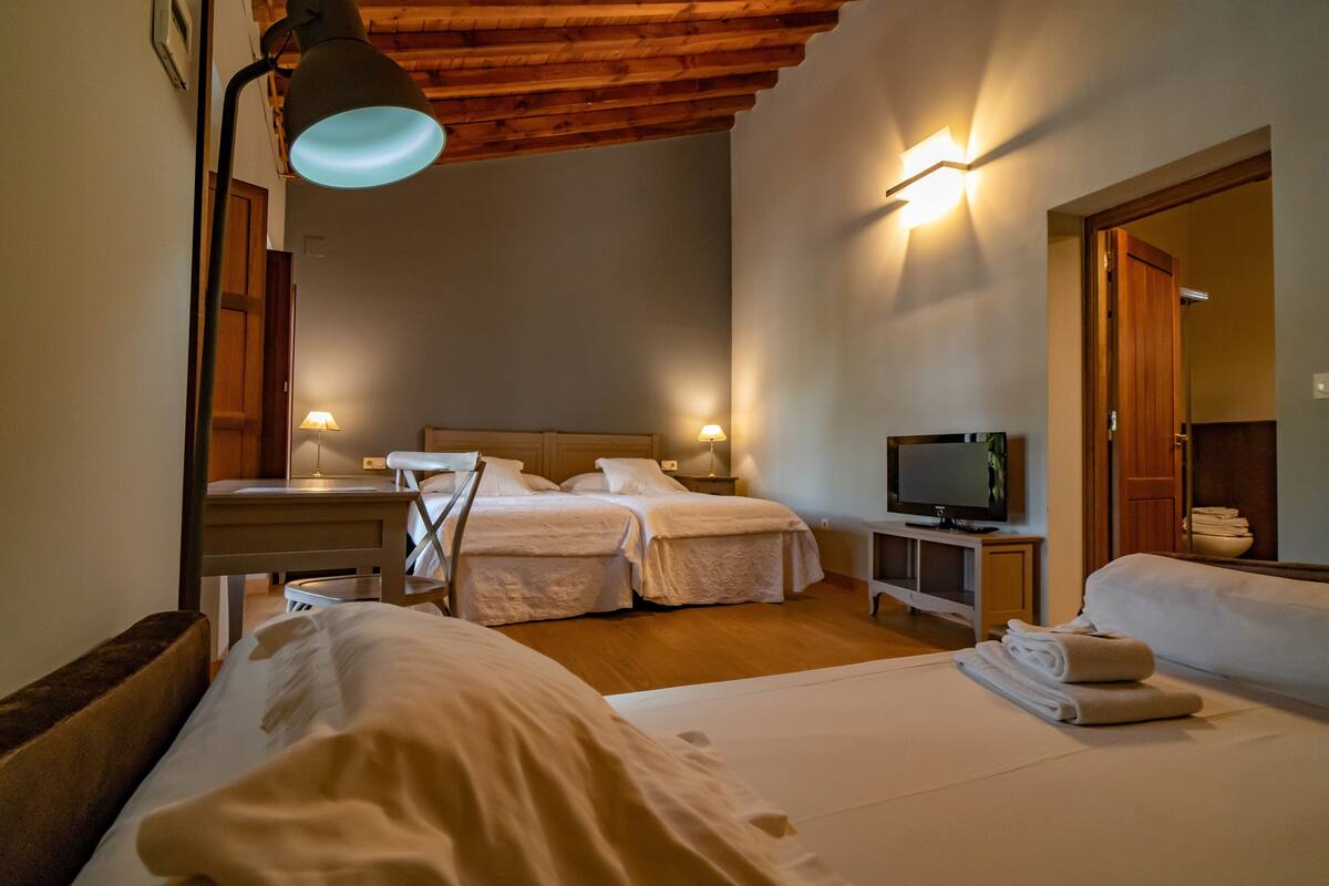 A hotel room with two beds, a television, a desk with a lamp, and a wooden ceiling.