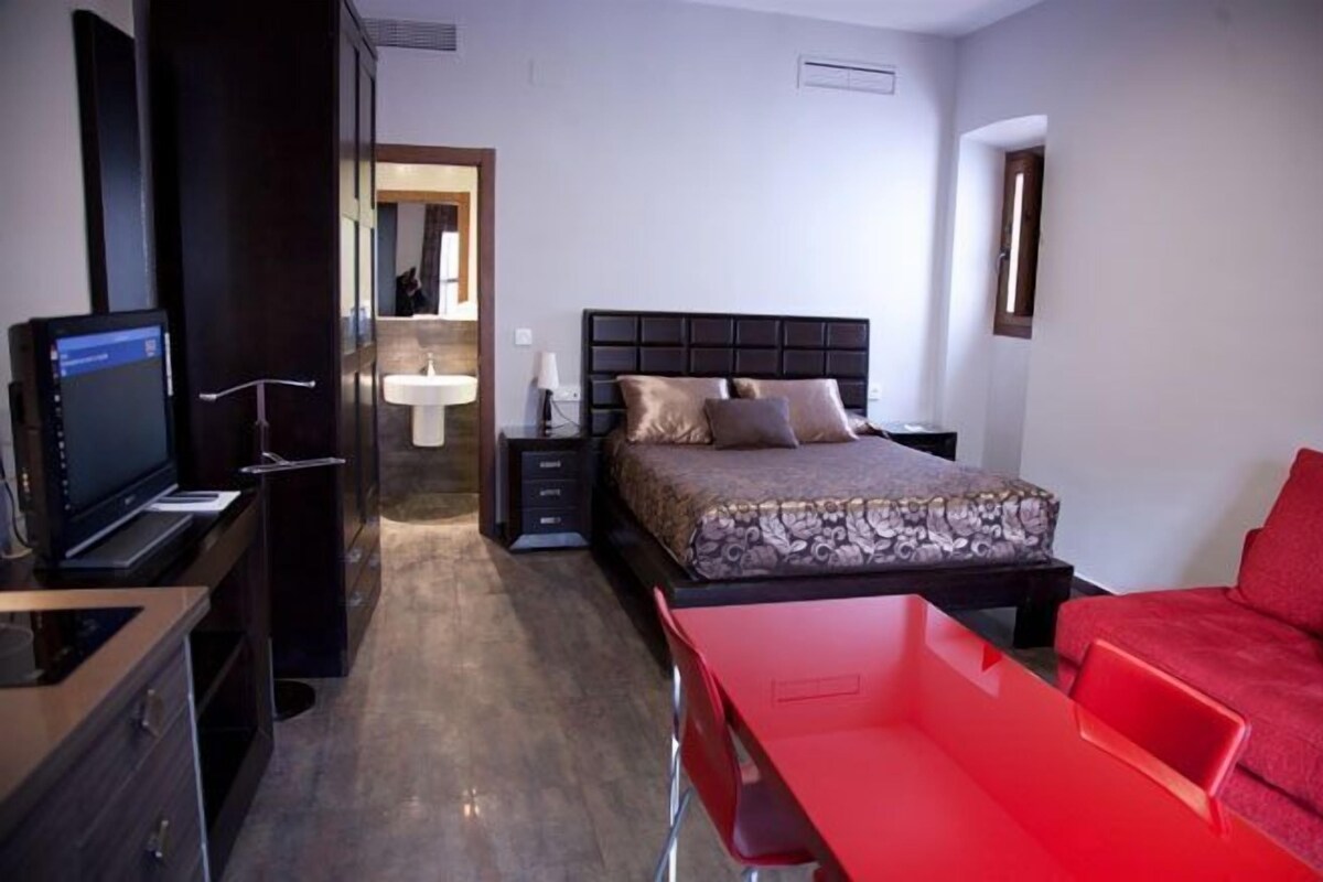 Quarto de hotel com uma cama, uma escrivaninha com um computador, uma cadeira vermelha e um sofá vermelho.