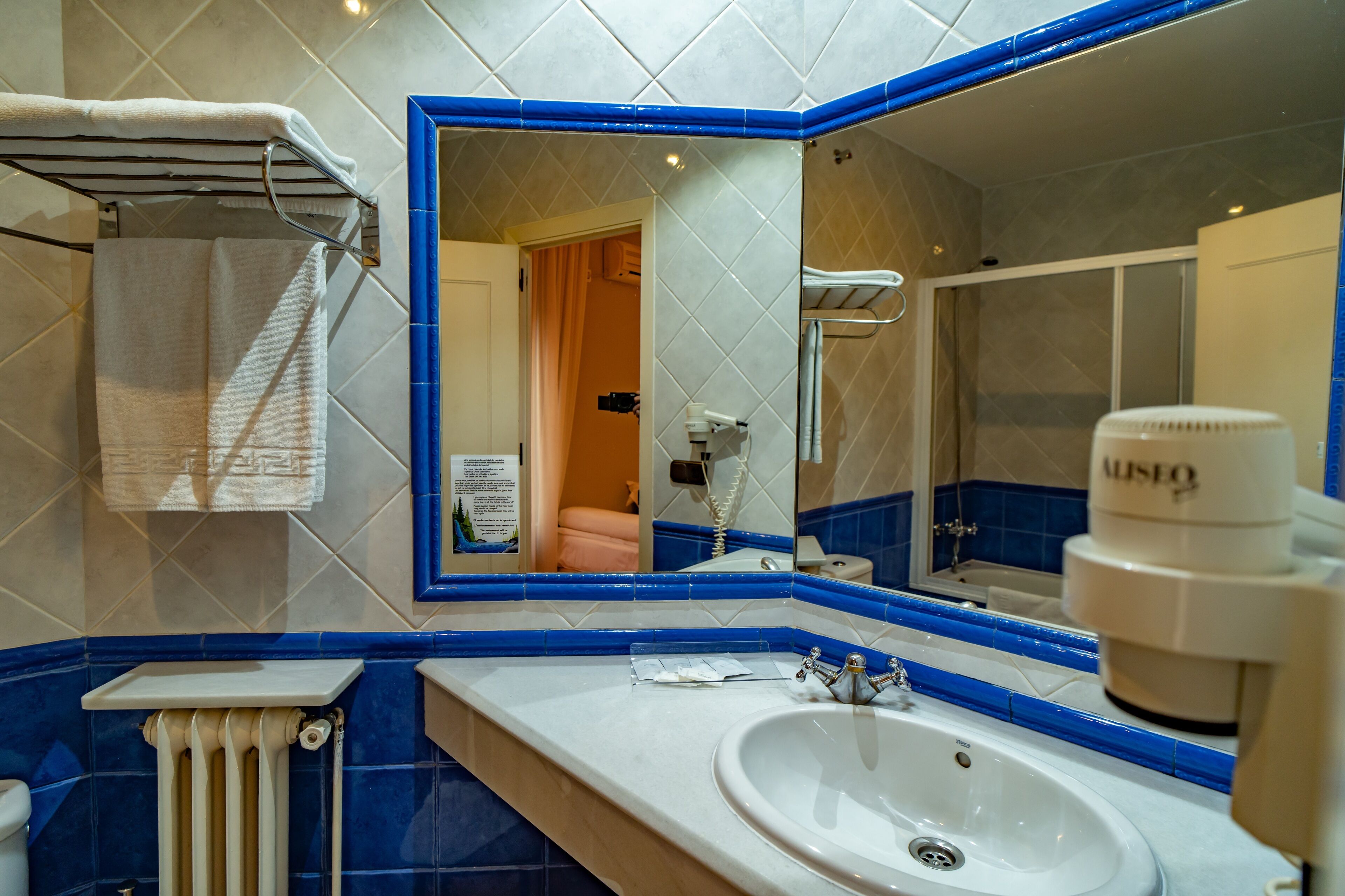 Une salle de bain avec un lavabo blanc, des murs recouverts de carreaux bleus, un grand miroir et un porte-serviettes.