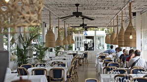Un restaurante con mesas dispuestas para comer, ventiladores de techo de mimbre y amplios ventanales.