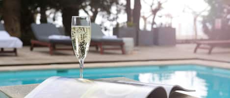 A glass of champagne on a stone ledge with a magazine, poolside chairs, and a pool in the background.