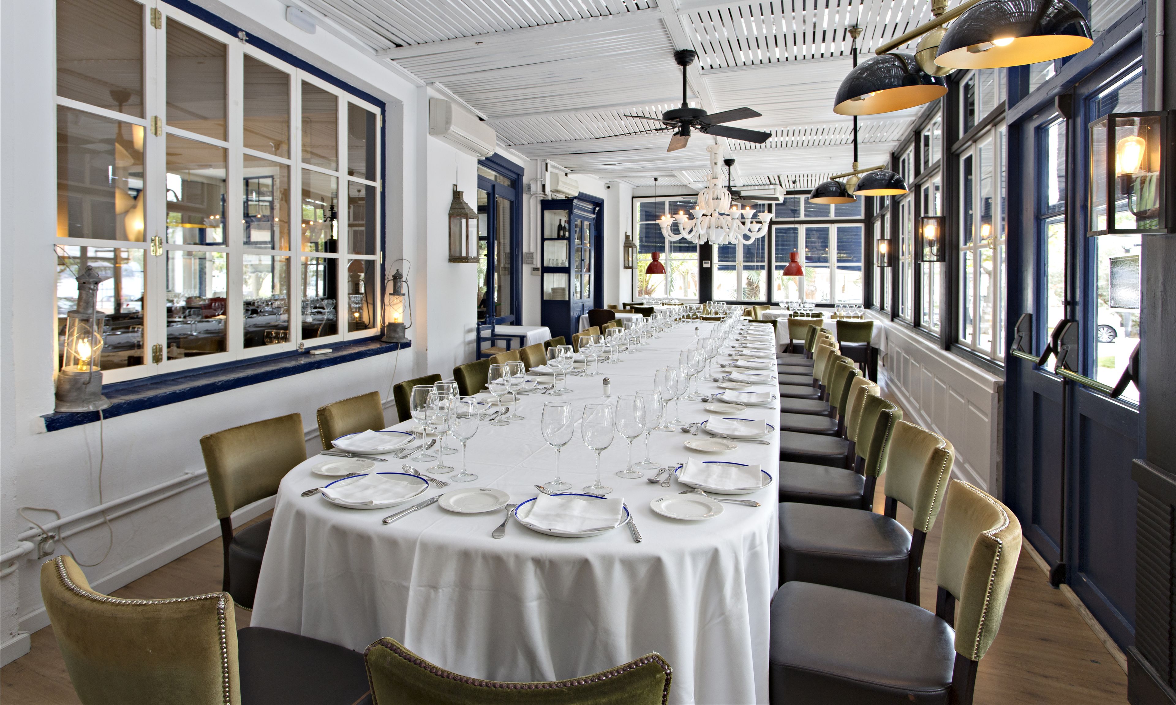 A long dining table set with white tablecloths, silverware, and wine glasses, surrounded by upholstered chairs.