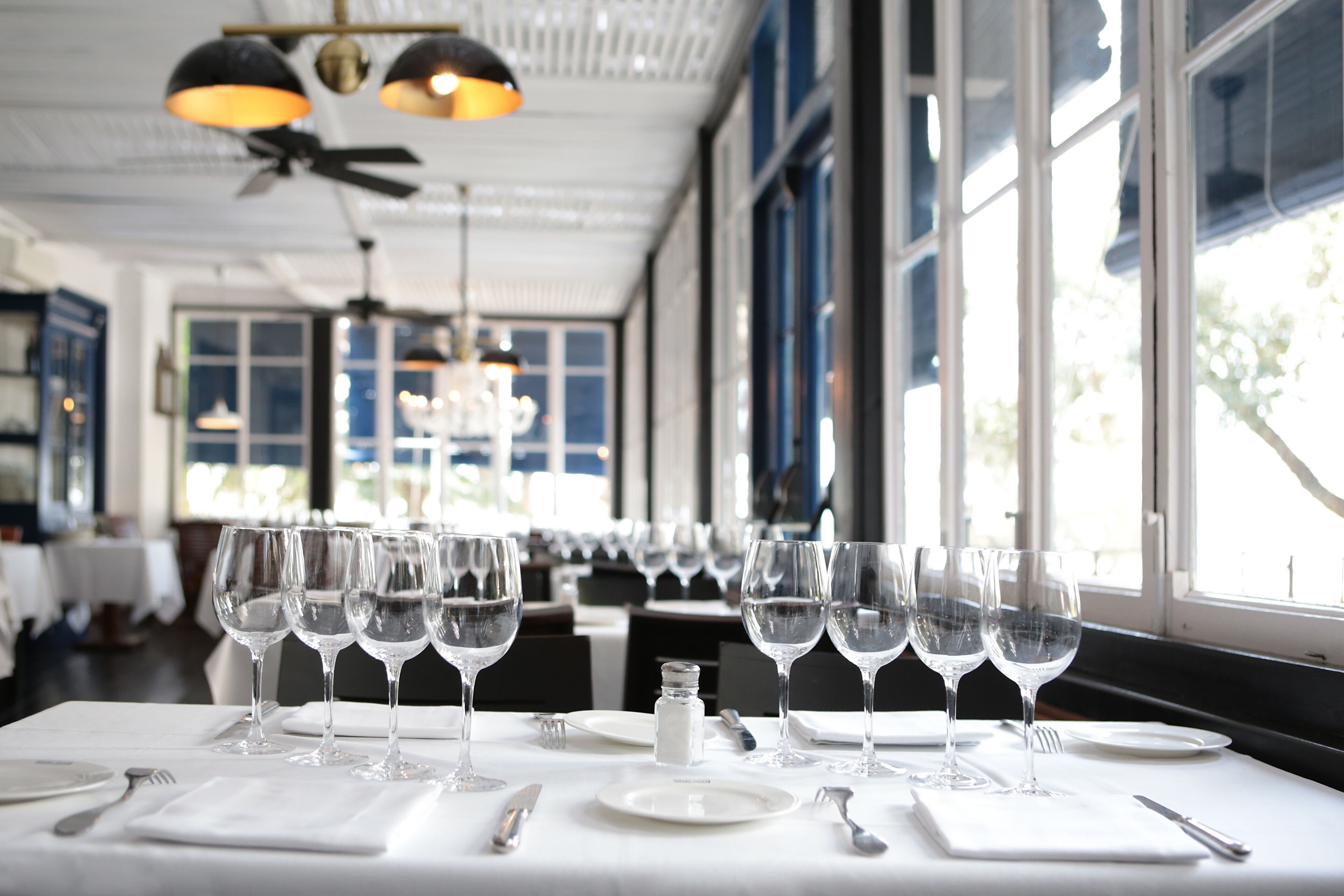 A dining area with multiple tables set for service, large windows, and ceiling fans.