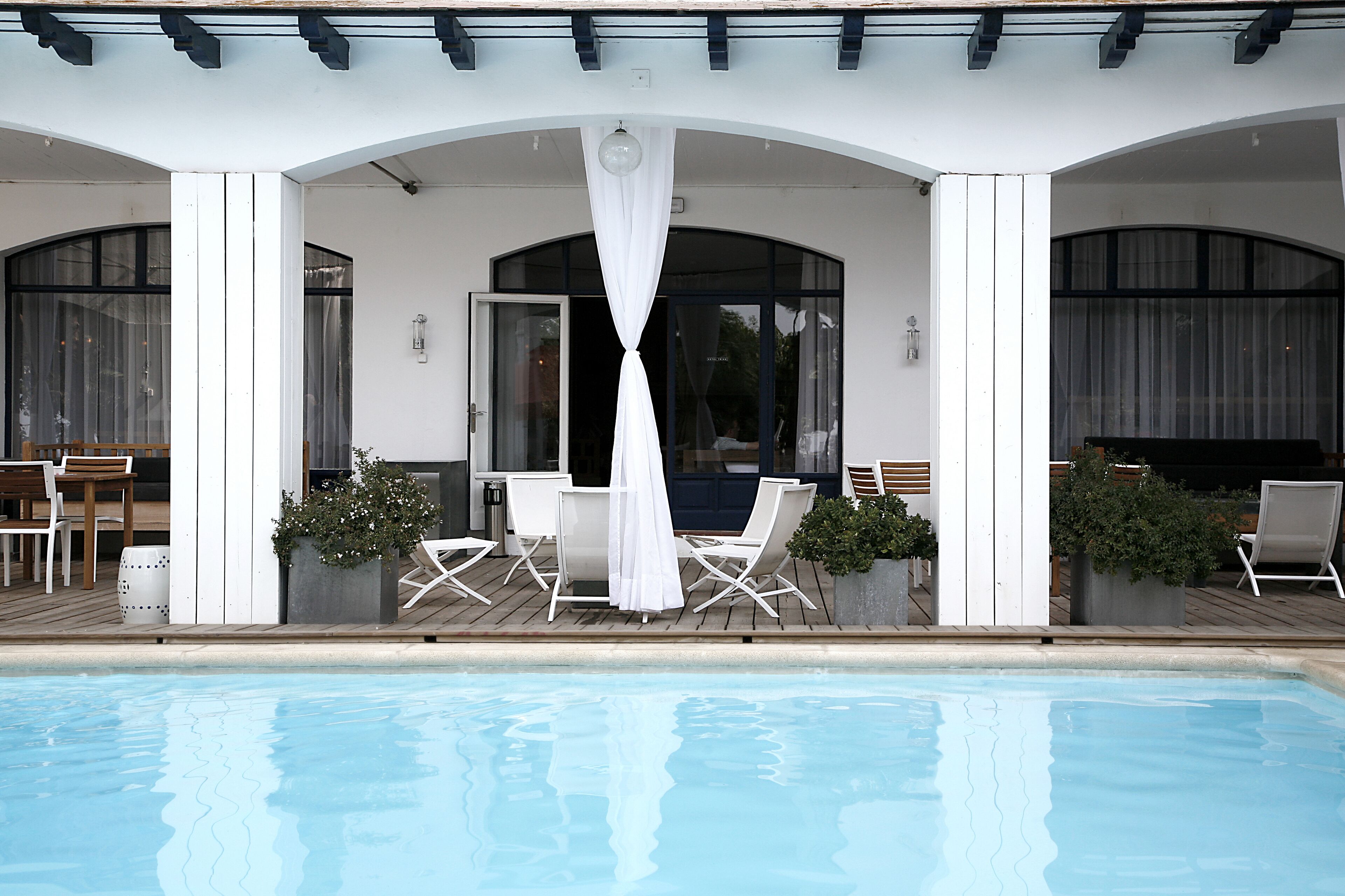 Un espace aménagé au bord de la piscine, avec un coin salon couvert, des chaises blanches et une piscine.