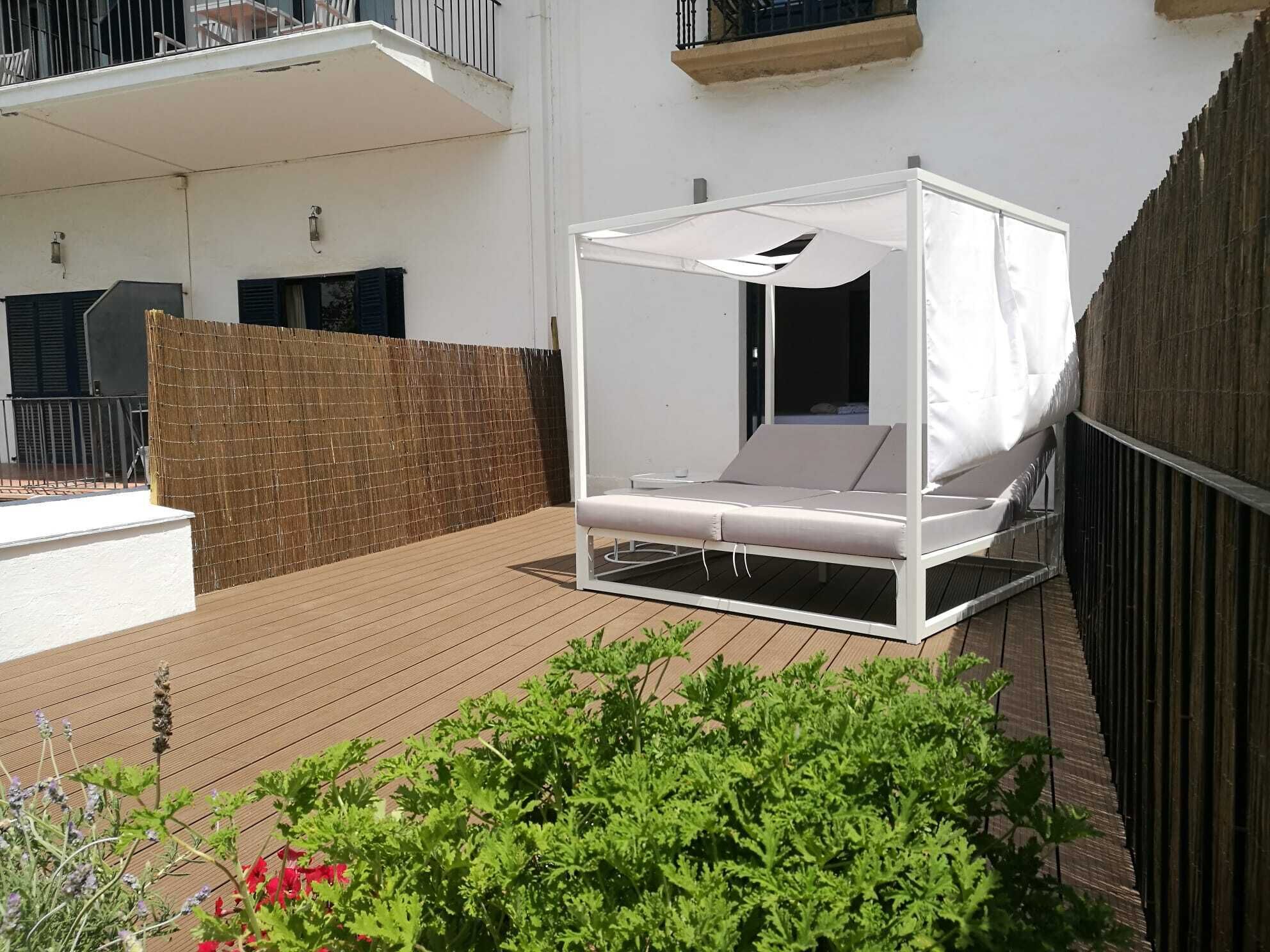 Una cama blanca al aire libre con dosel, rodeada por una terraza de madera y una cerca de bambú.