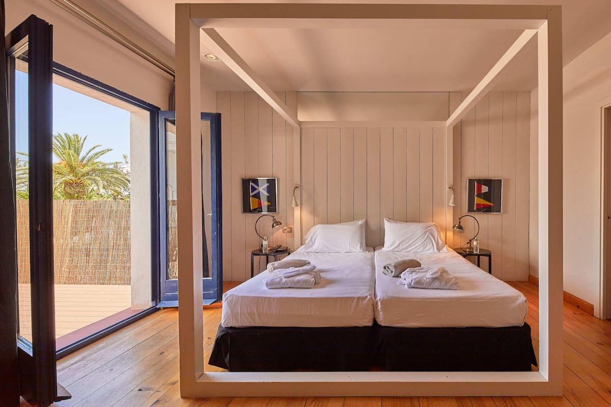 A double bed with white linens and towels, a small bedside table with a lamp, and a view of palm trees through a glass door.