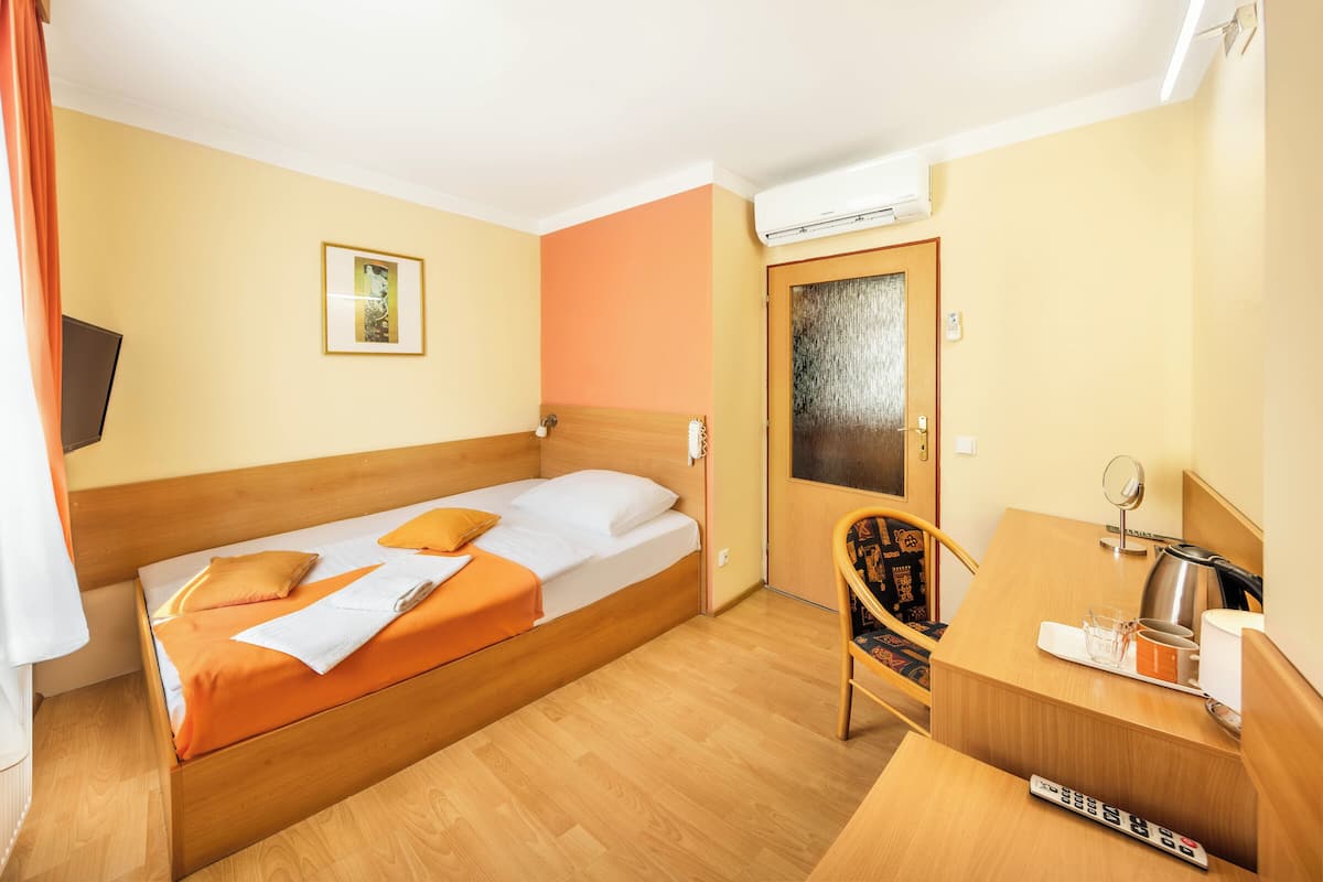 A hotel room with a single bed, a desk with a chair, a small table, a mirror, and an air conditioning unit.
