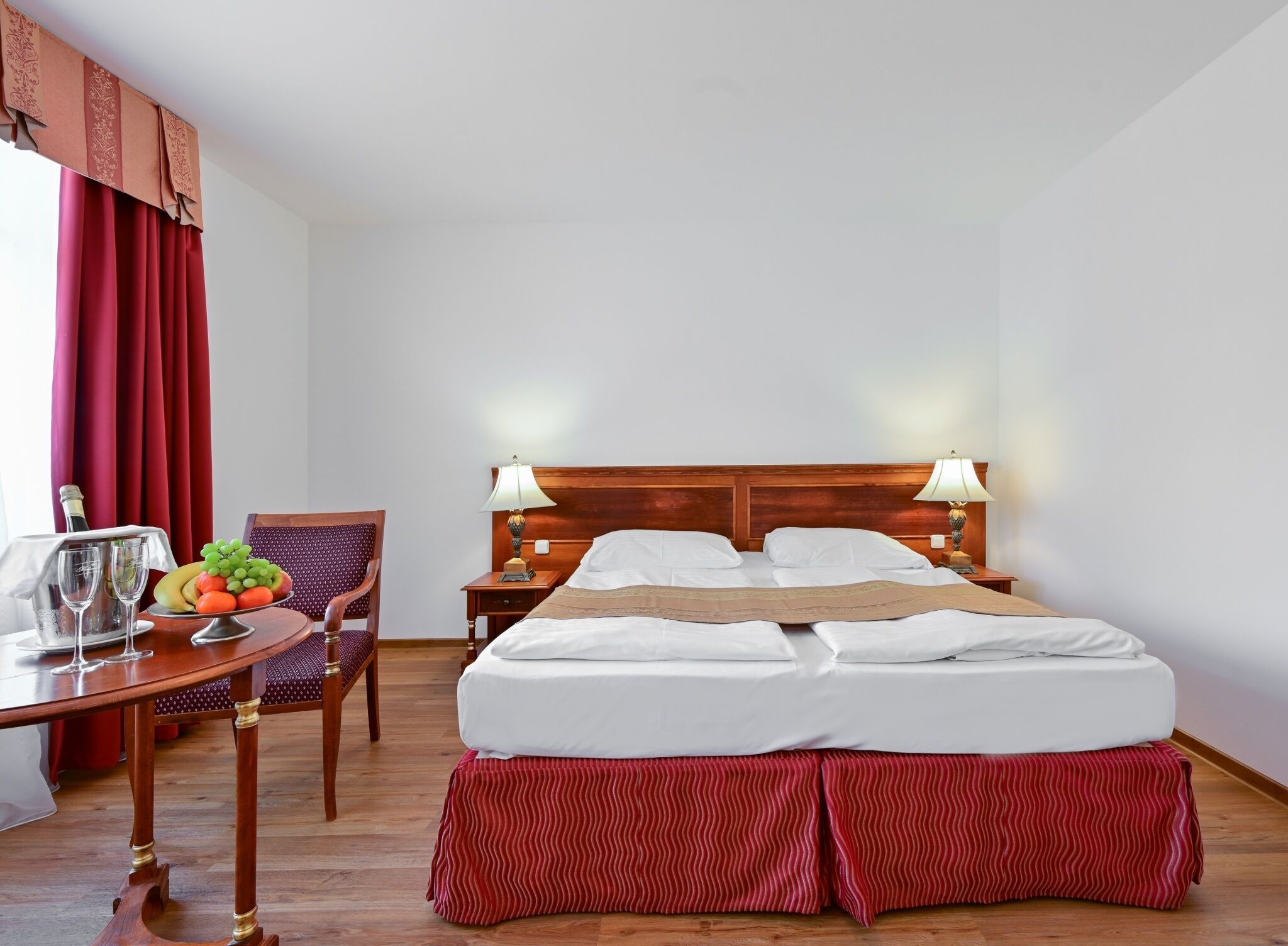 Een hotelkamer met een bed, bedlampen, een houten nachtkastje, een stoel, een tafel met een schaal fruit en glazen, en een rood gordijn.