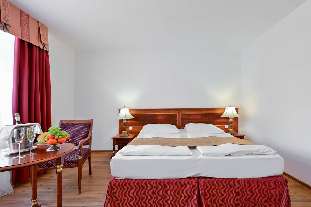 A hotel room with a bed, bedside lamps, a wooden nightstand, a chair, a table with a fruit bowl and glasses, and a red curtain.