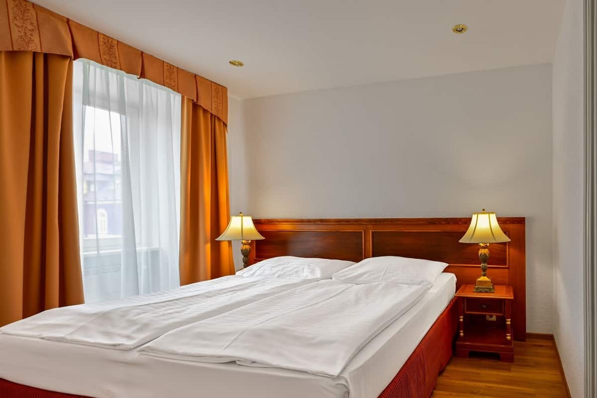 A hotel room with a bed, two bedside lamps, a nightstand, and a window with curtains.