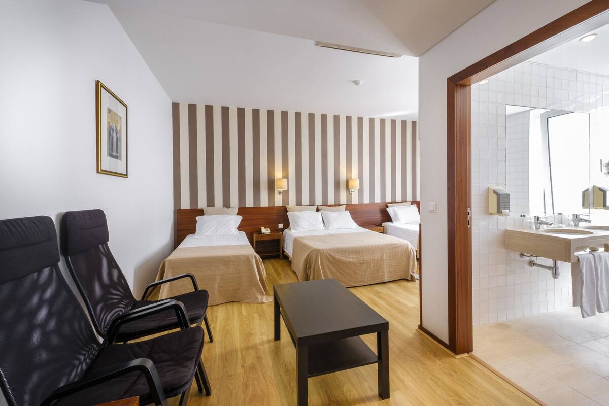A hotel room with two beds, a wooden headboard, a small table, a chair, a bathroom with a sink and mirror, and a striped wall.