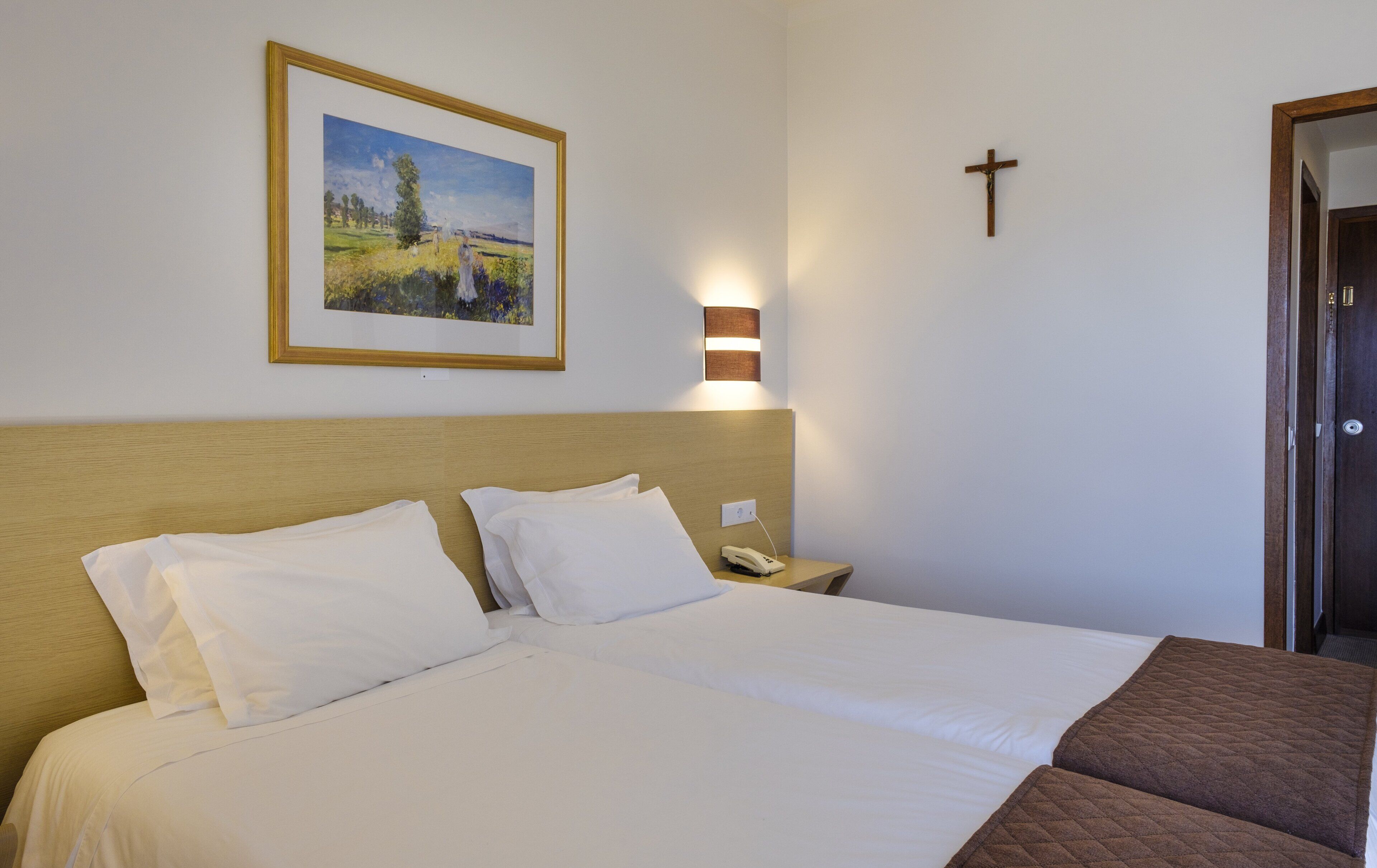 Une chambre d’hôtel avec un lit, une table de chevet, un crucifix fixé au mur et un tableau encadré.