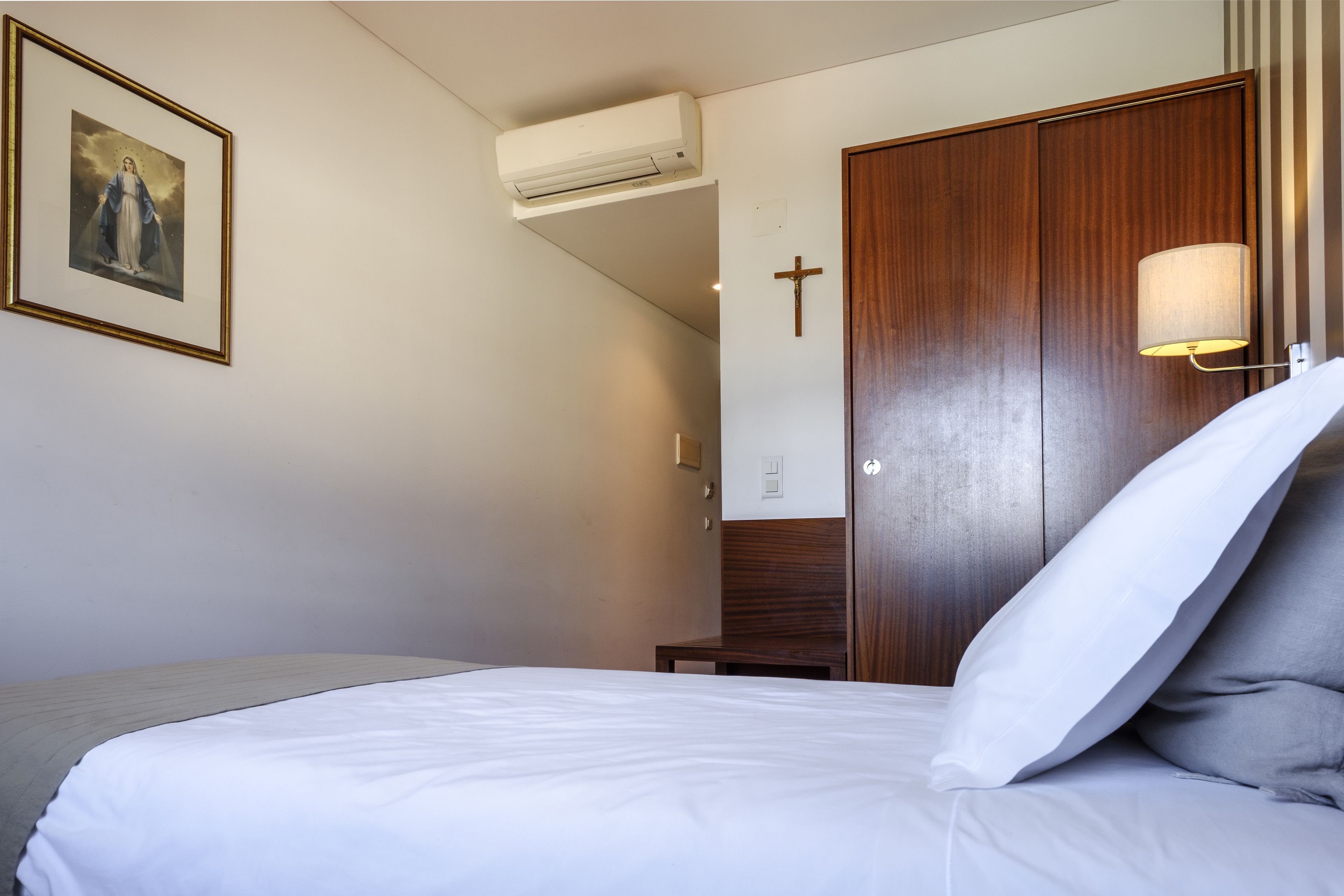 A hotel room with a bed, a wooden wardrobe, a cross on the wall, and a framed picture above the bed.
