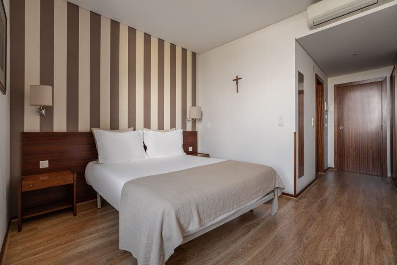 Habitación de hotel con una cama grande, cabecero de madera, dos mesitas de noche, una cruz en la pared y un equipo de aire acondicionado.