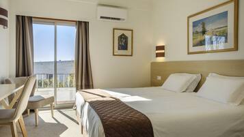 Habitación de hotel con cama, escritorio y una silla. Cuenta con un ventanal con cortinas, aire acondicionado y dos cuadros en la pared.