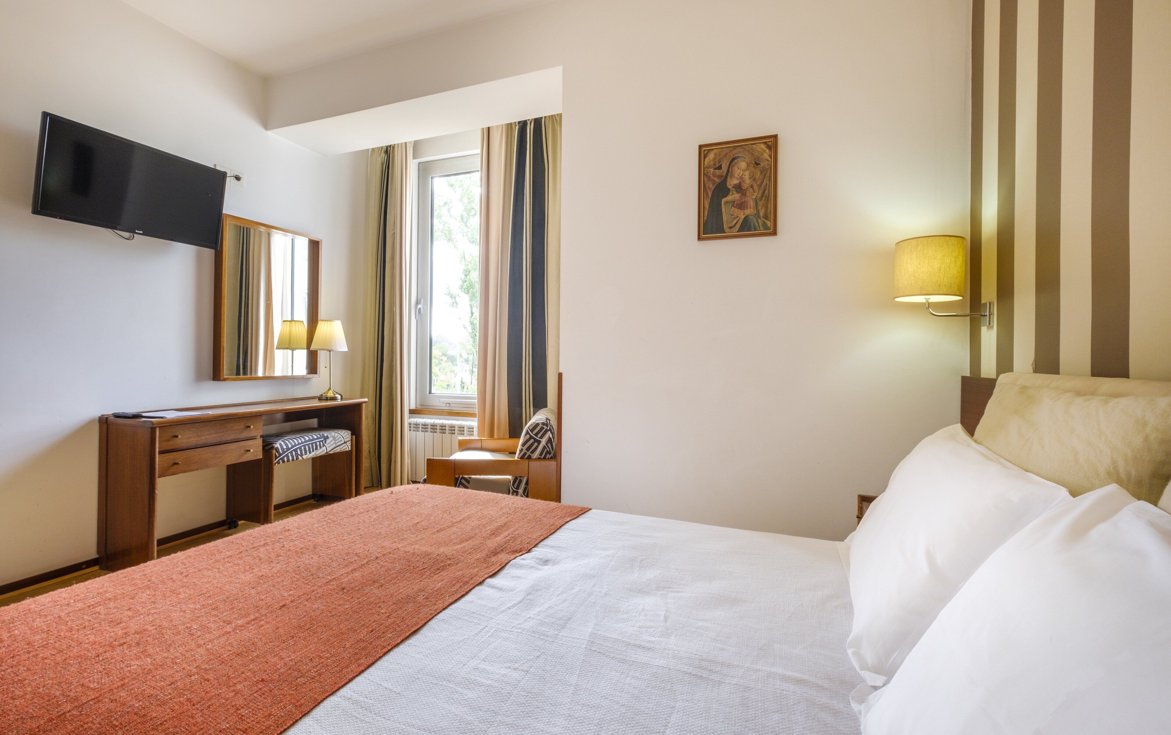 Una habitación de hotel con cama, escritorio, televisor, un cuadro y ventana con cortinas.