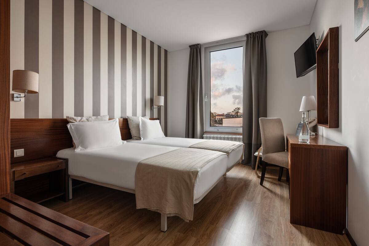 A hotel room with two beds, a desk, a chair, a TV, and a window with curtains.