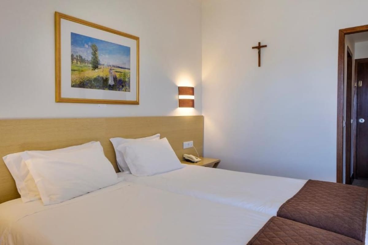 Habitación de hotel con una cama grande, una mesita de noche con teléfono, un cuadro con paisaje enmarco, una lámpara de pared y una cruz de madera.