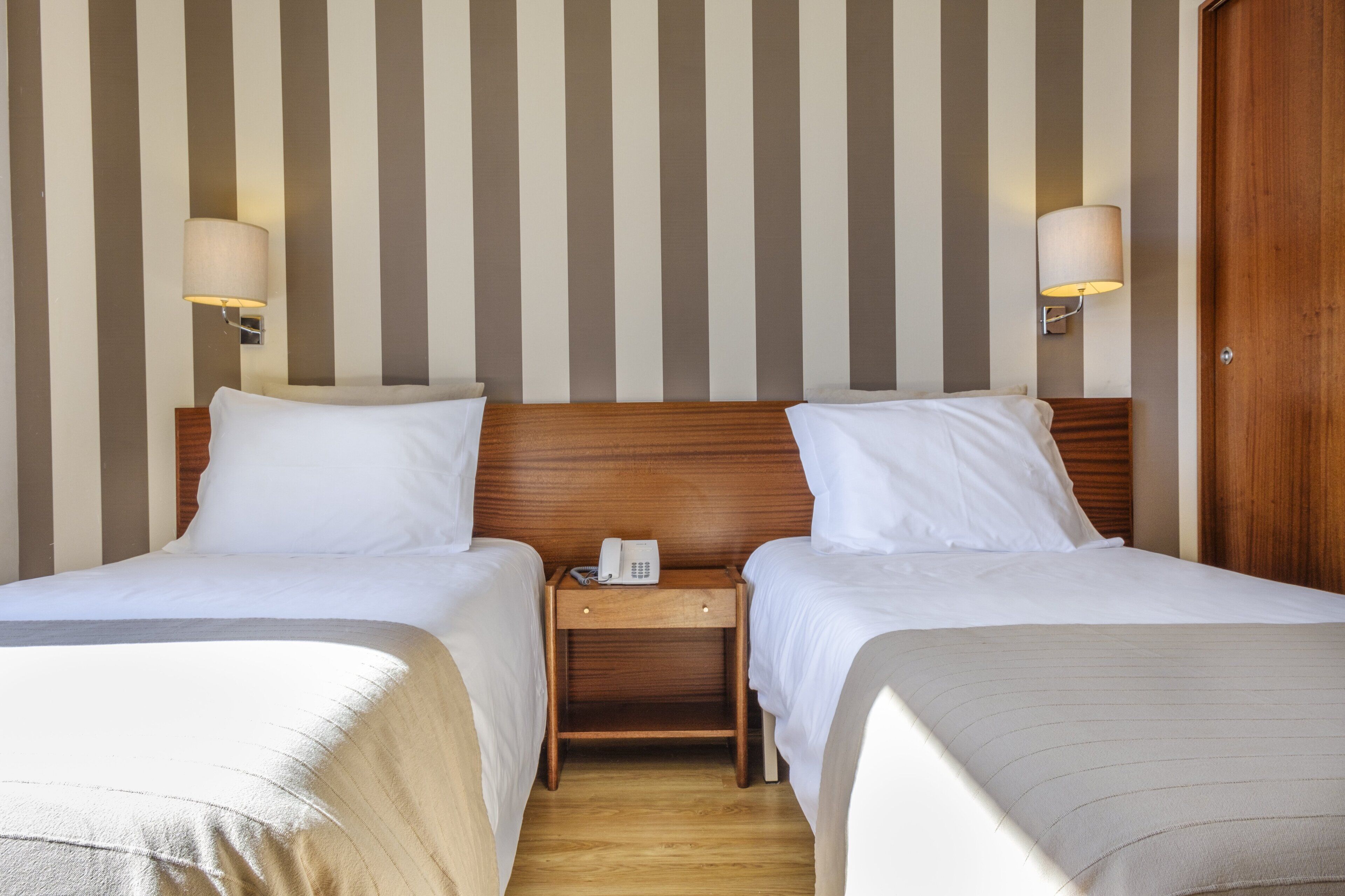 A hotel room with two beds, a wooden nightstand with a phone, and wall-mounted lamps.