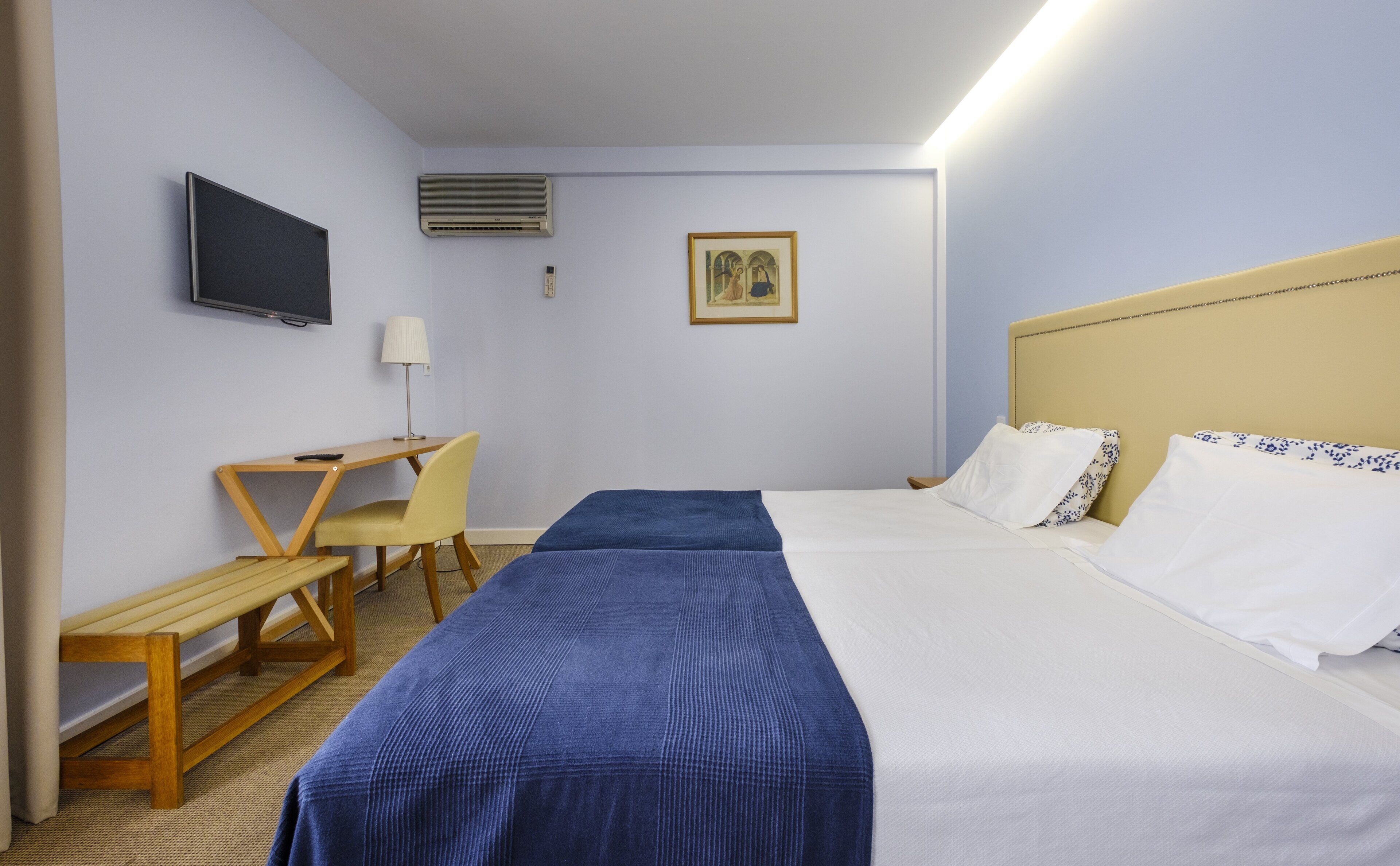 Habitación de hotel con cama, escritorio, silla, televisión, aire acondicionado y un cuadro en la pared.