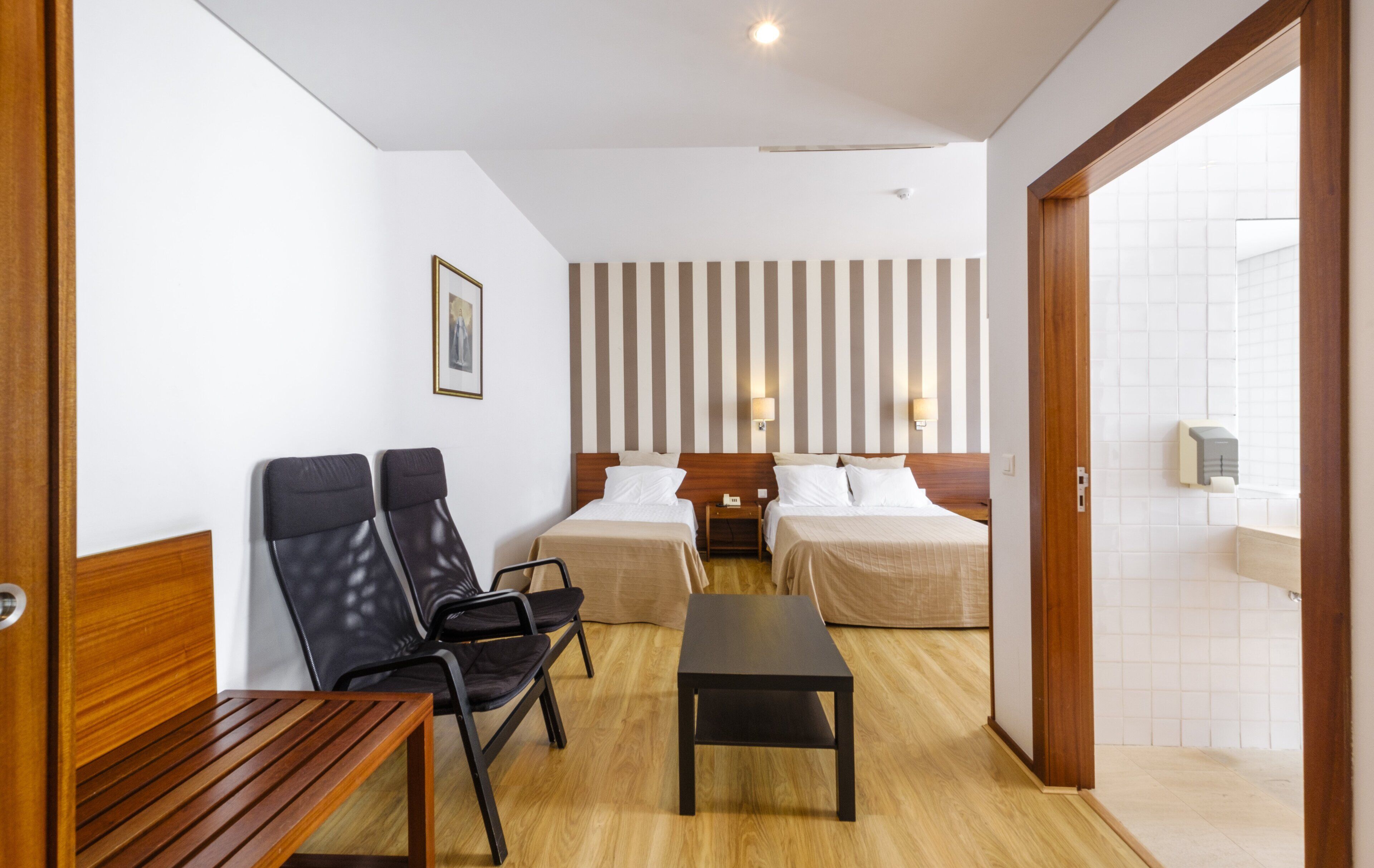 A hotel room with two beds, a wooden bench, a small table, and a chair.