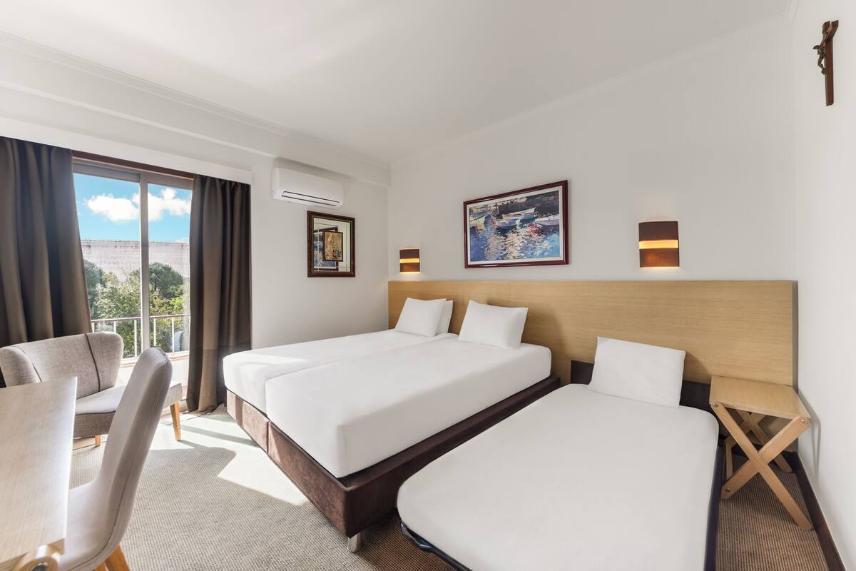 A hotel room with two beds, a sofa, a desk, and a large window with curtains.