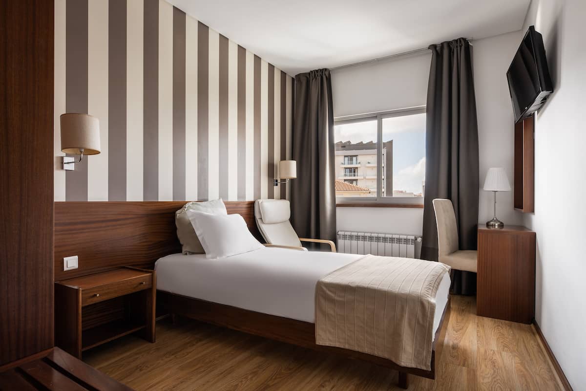 A hotel room with a bed, two chairs, a desk, a TV, and a window with curtains.