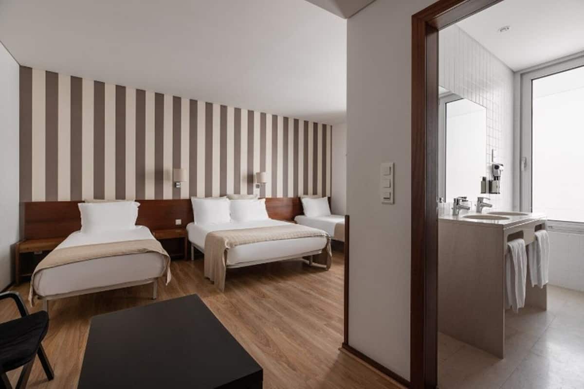 A hotel room with two beds, a small table, a chair, a bathroom with a sink and mirror, and a striped wall.