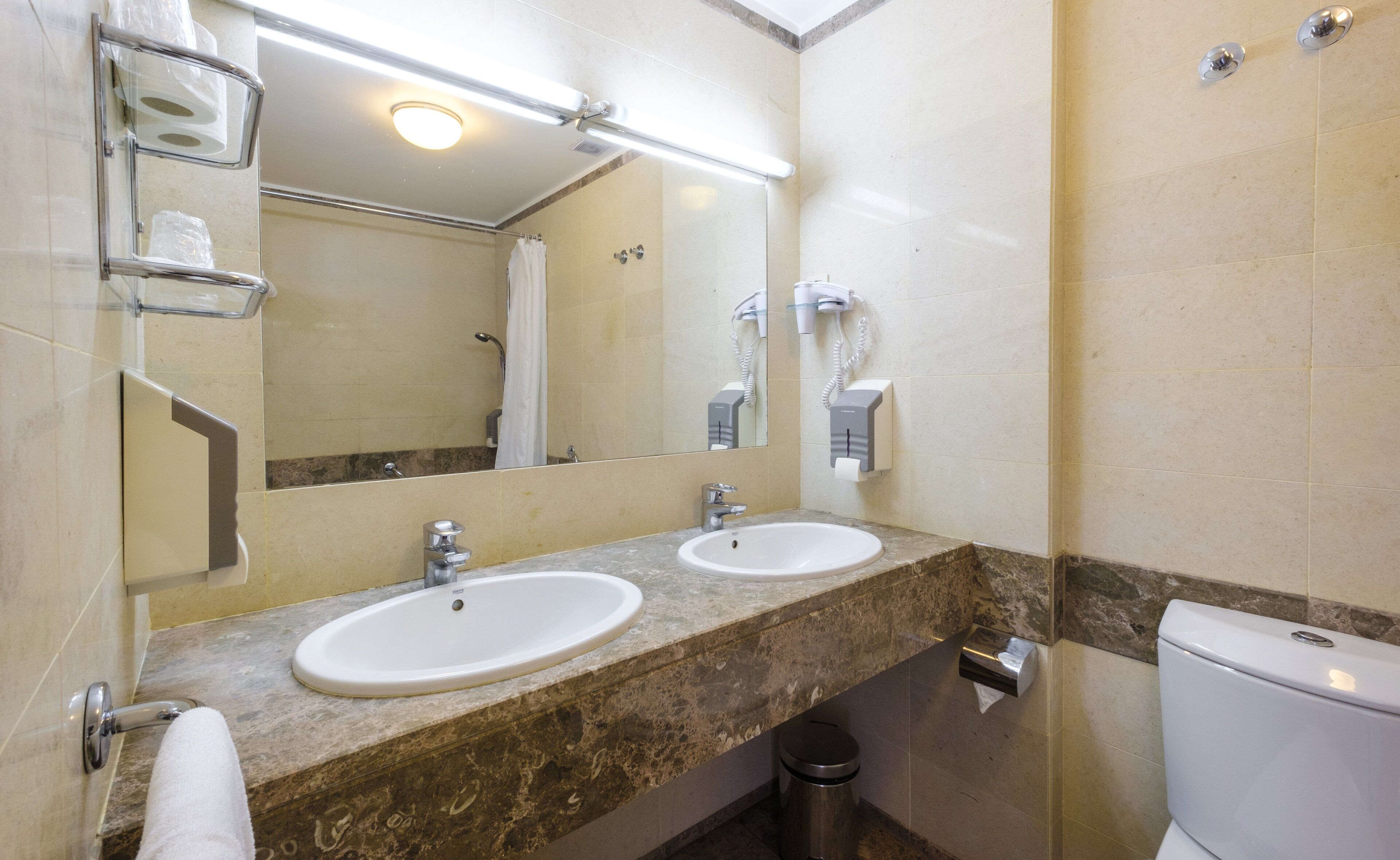A bathroom with two sinks, a mirror, a toilet, and a trash can.