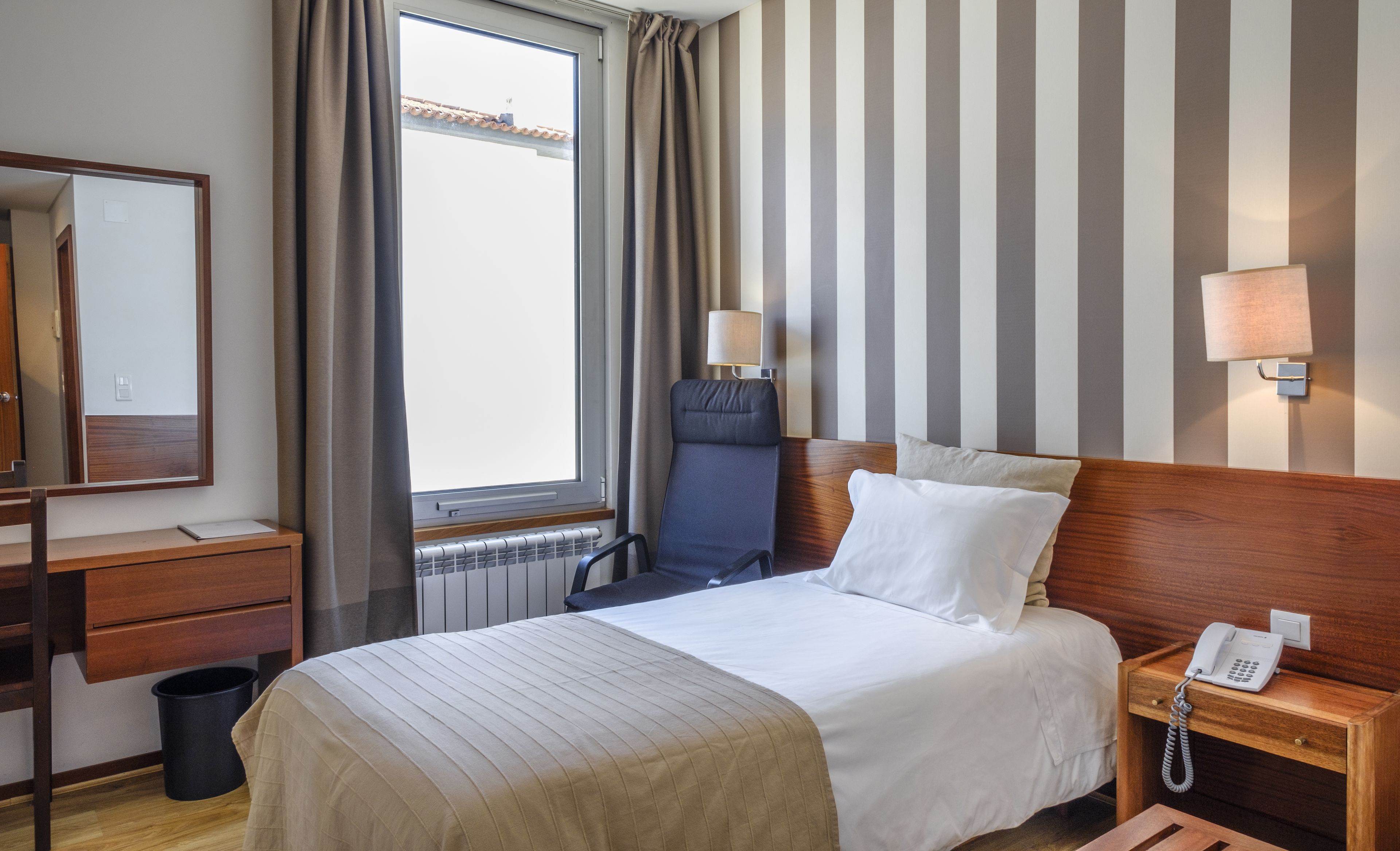 A hotel room with a bed, a desk, a chair, a lamp, and a telephone.
