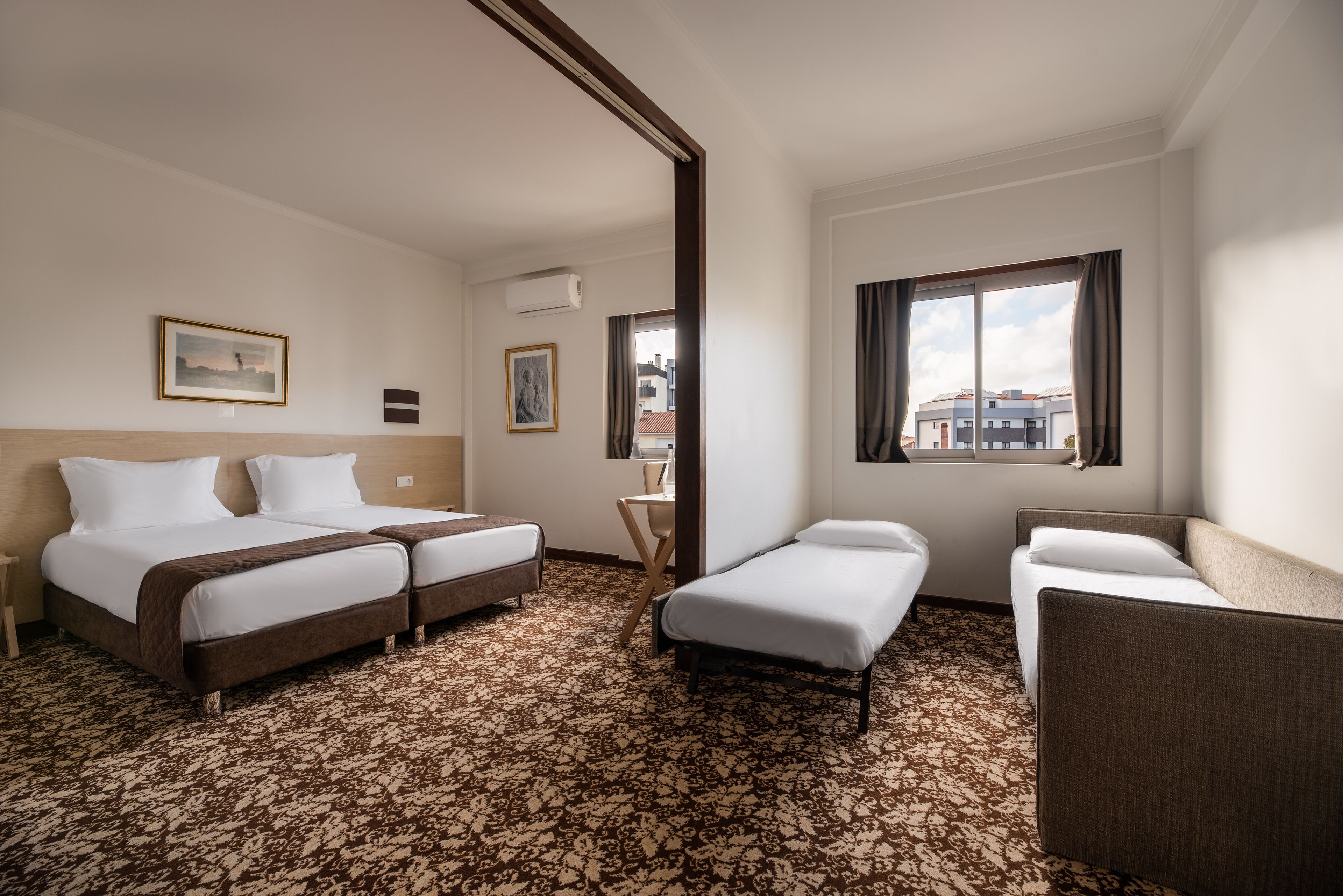 A hotel room with two beds, a small sofa, a window with curtains, and a patterned carpet.