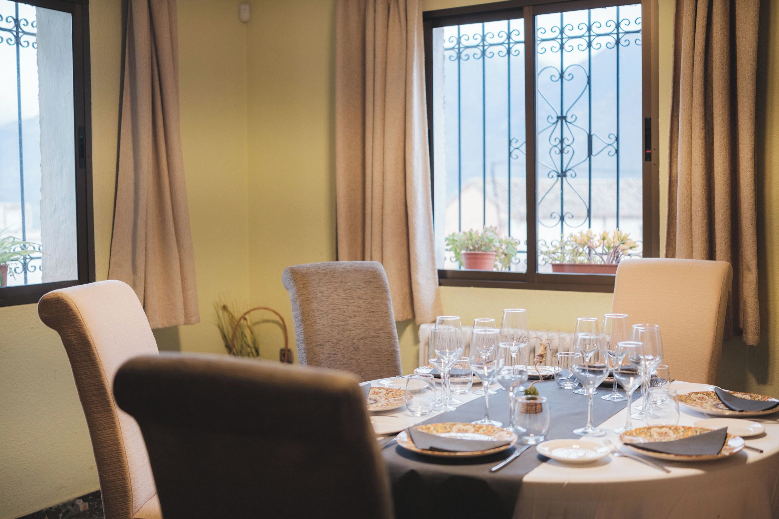 Mesa lista para una comida, con sillas, una ventana con cortinas y vistas al exterior.