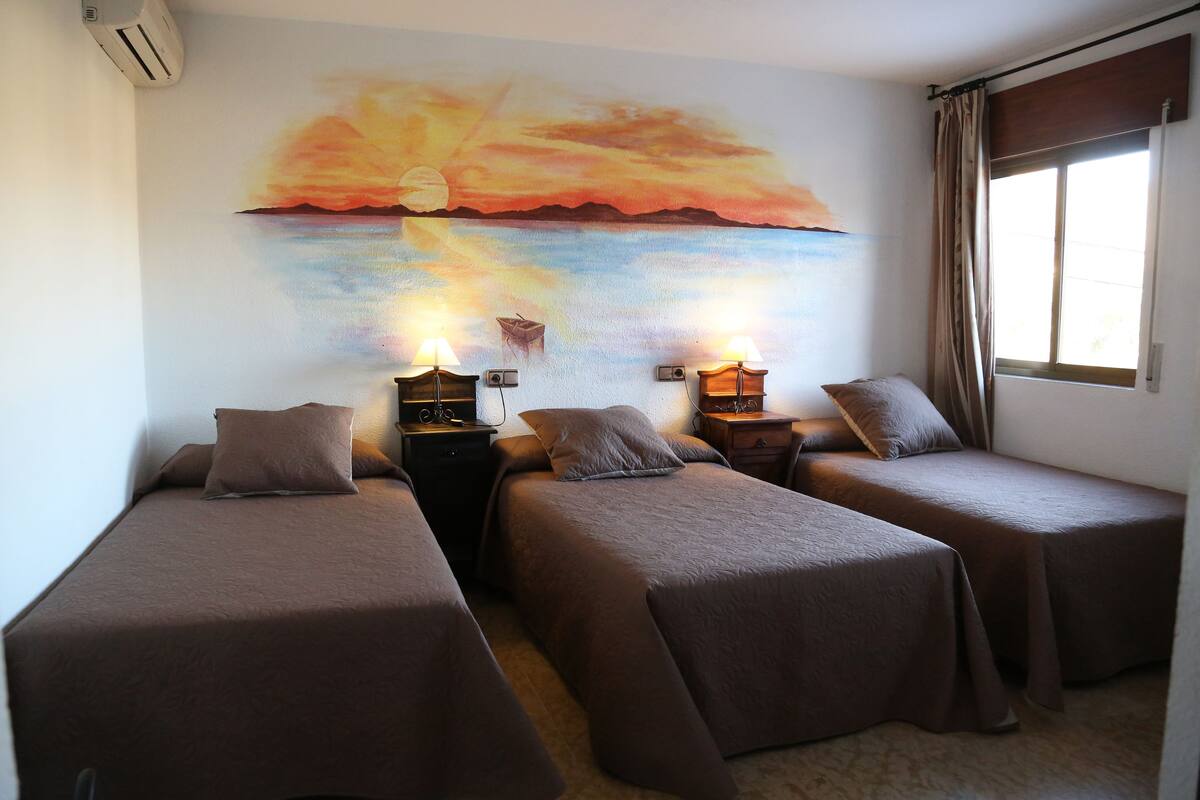 A room with two single beds, a nightstand with a lamp, a wall painting of a sunset, and a window with curtains.