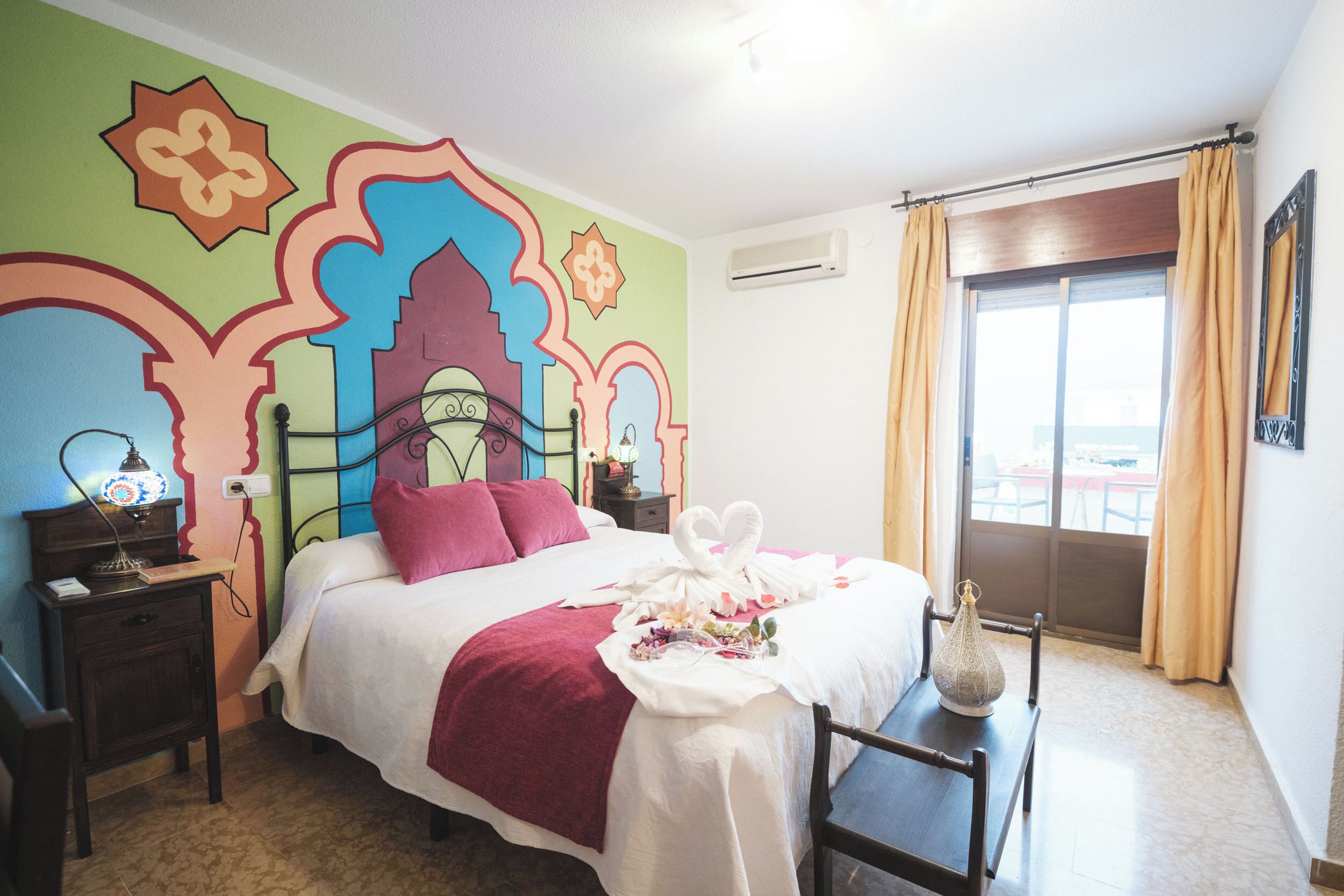 Un llit ben fet amb llençols vermells, una taula de nit amb una làmpada i un mural a la paret.