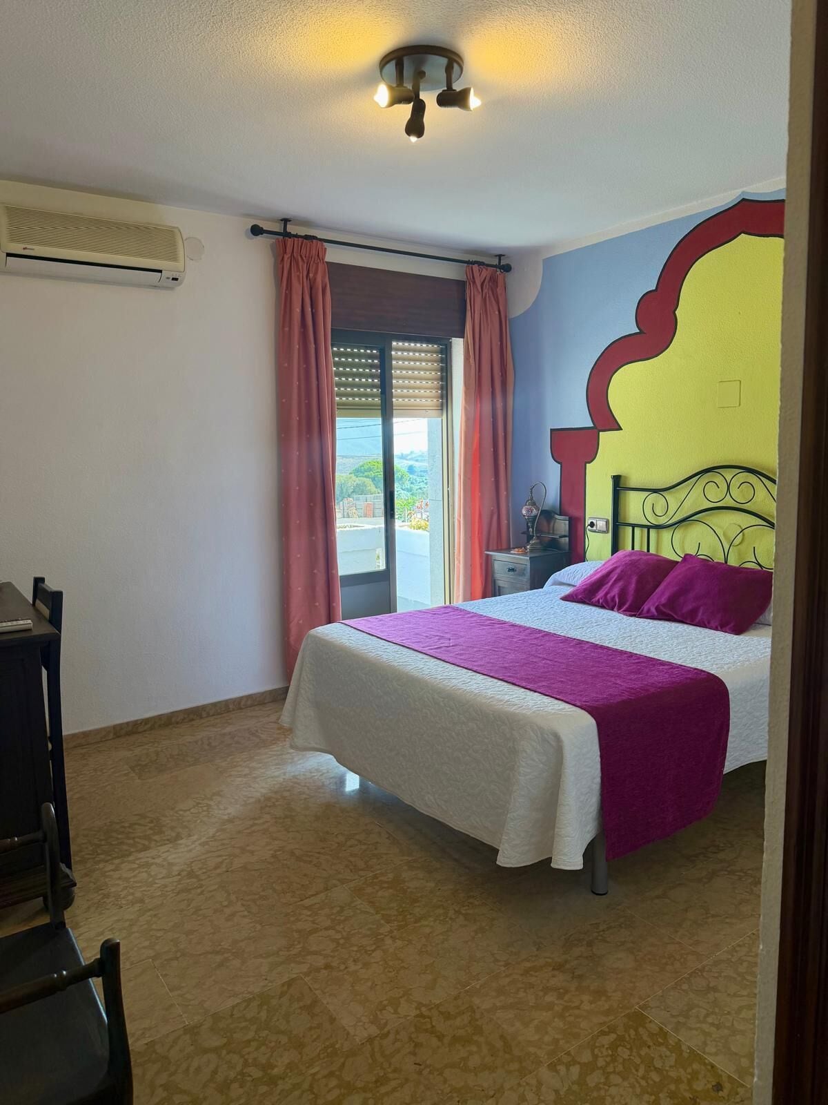 A bedroom with a bed, a nightstand, a window with curtains, and an air conditioning unit.