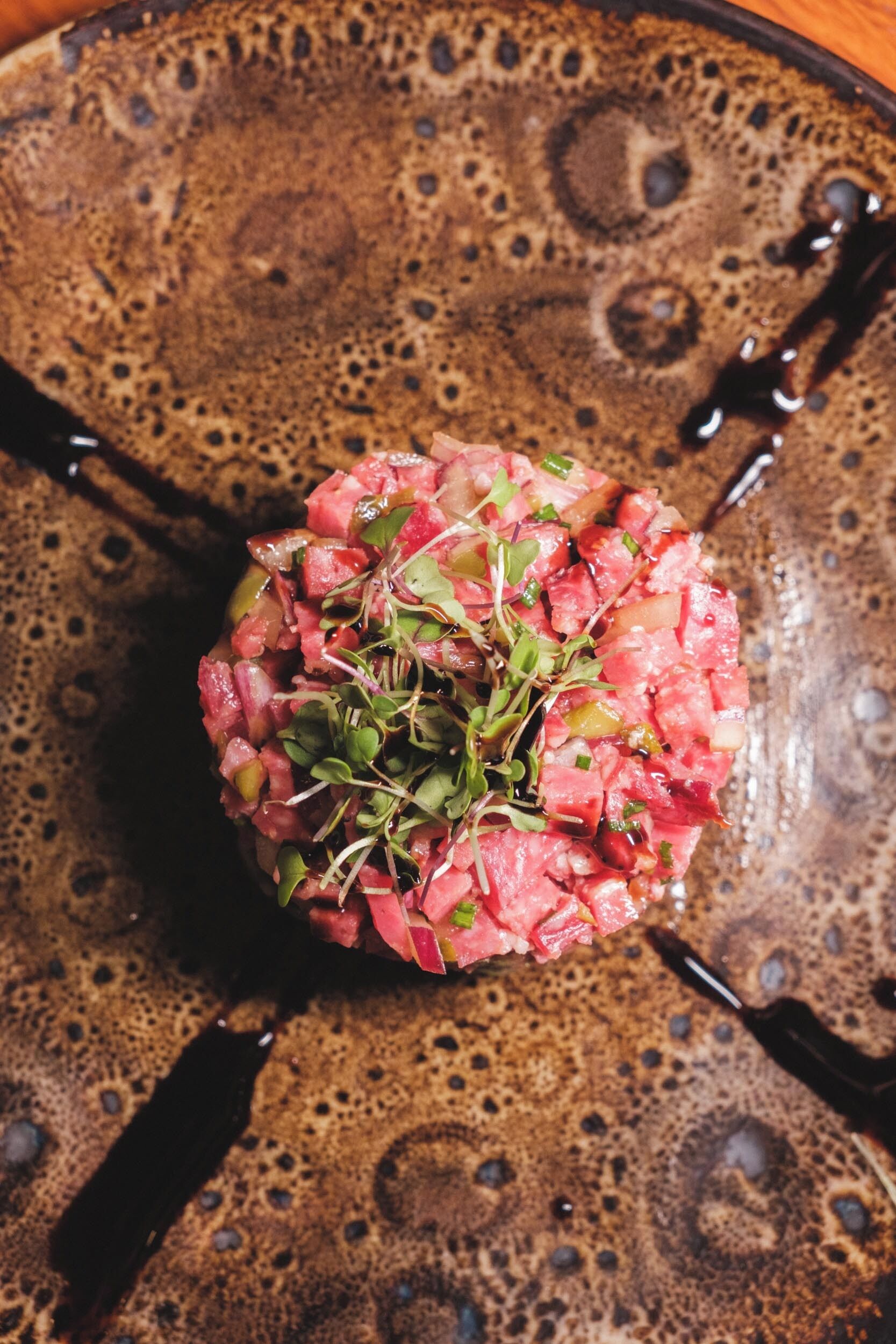 A round, textured plate with a central arrangement of diced pink meat and green garnish, surrounded by a dark sauce.