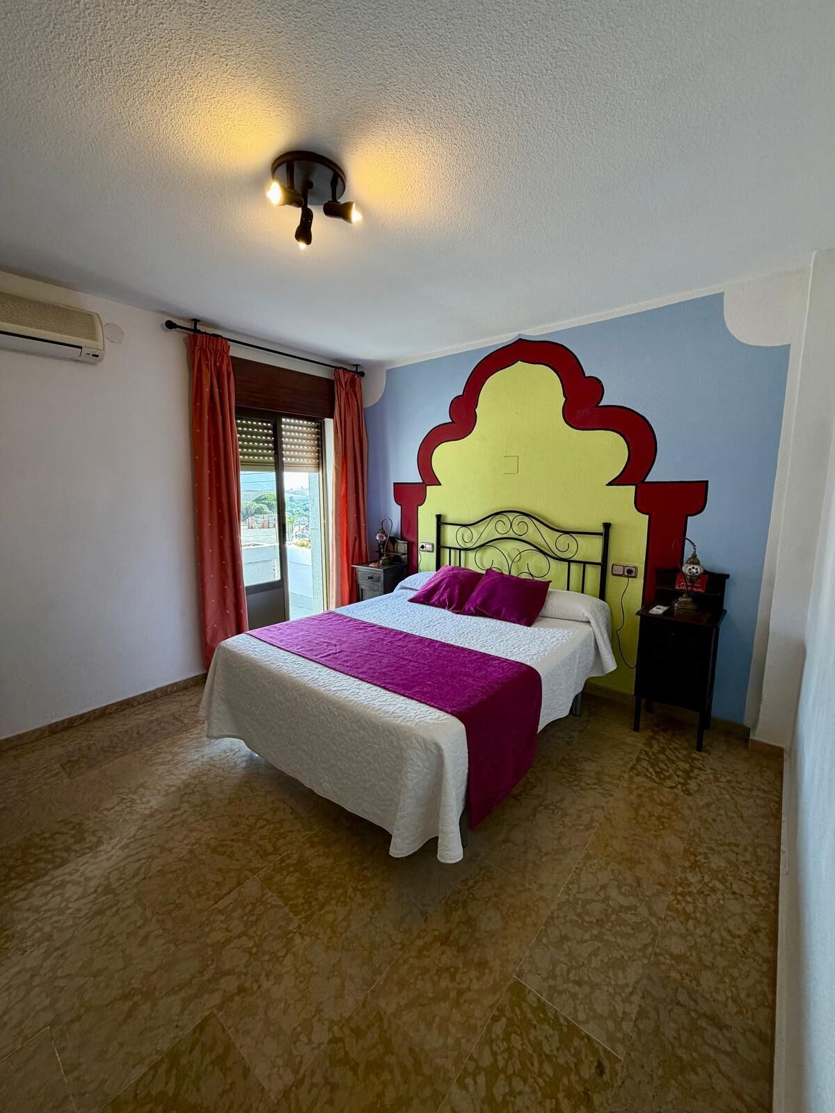 A bedroom with a bed, a nightstand, a wall-mounted light fixture, and a window with curtains.