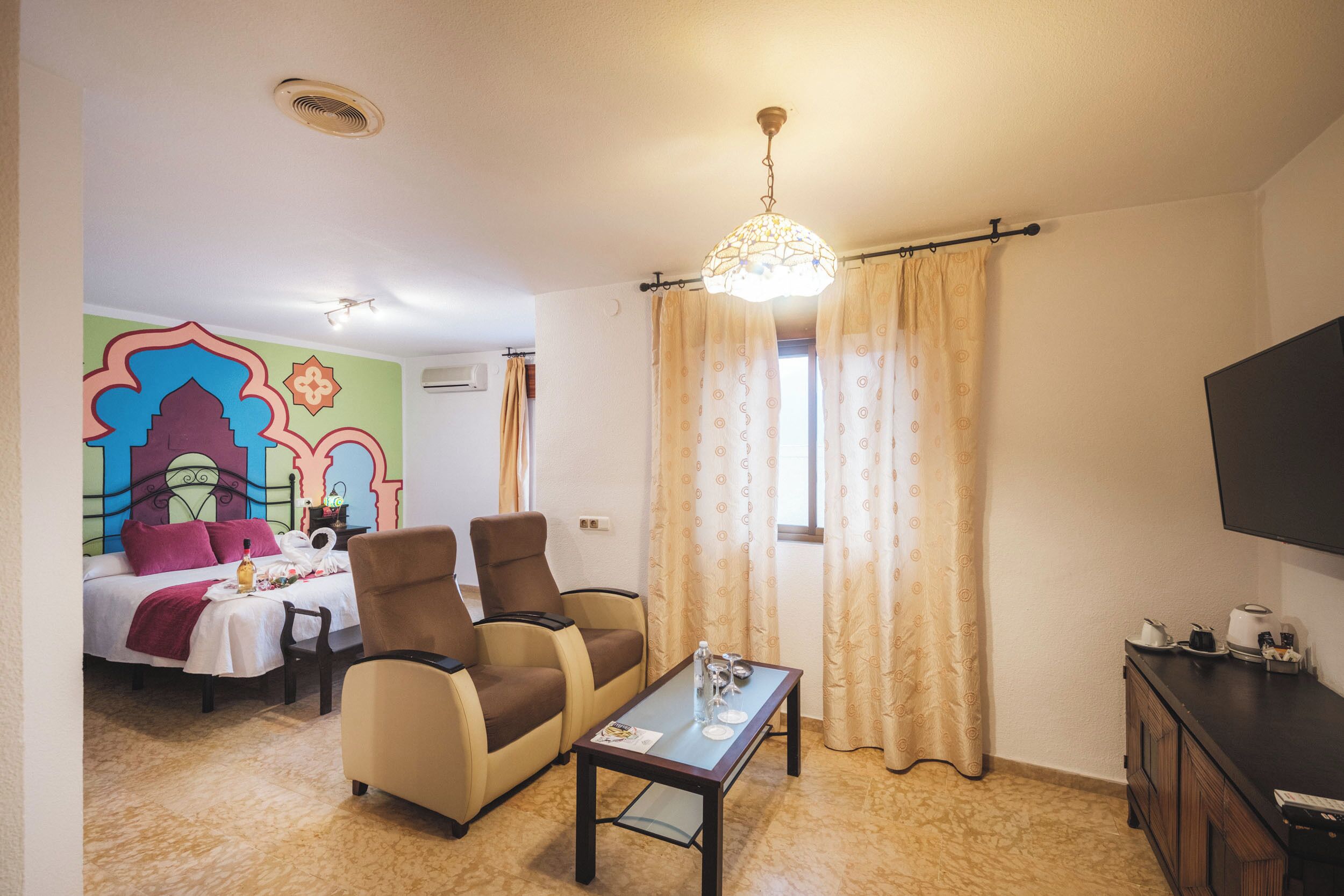 A hotel room with a bed, two armchairs, a small table, a TV, and a window with curtains.