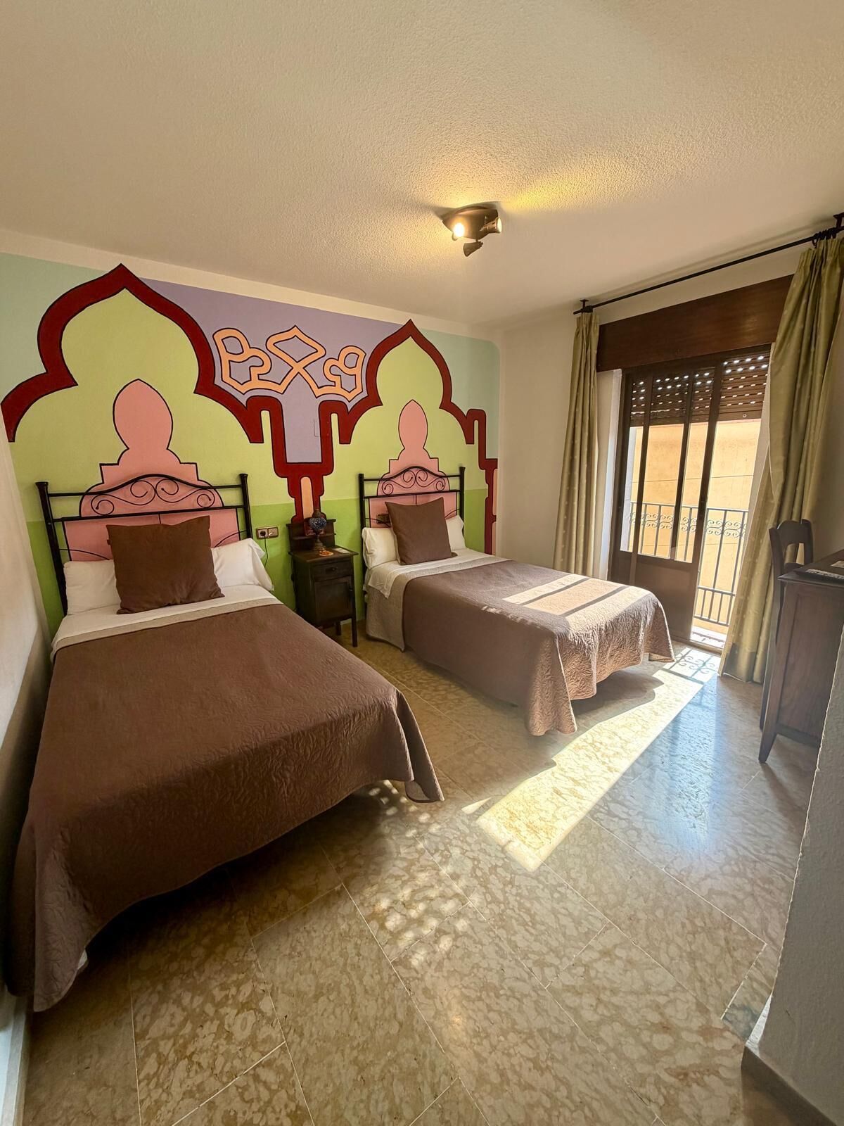 A hotel room with two beds, a decorative wall mural, a window with curtains, and a balcony.